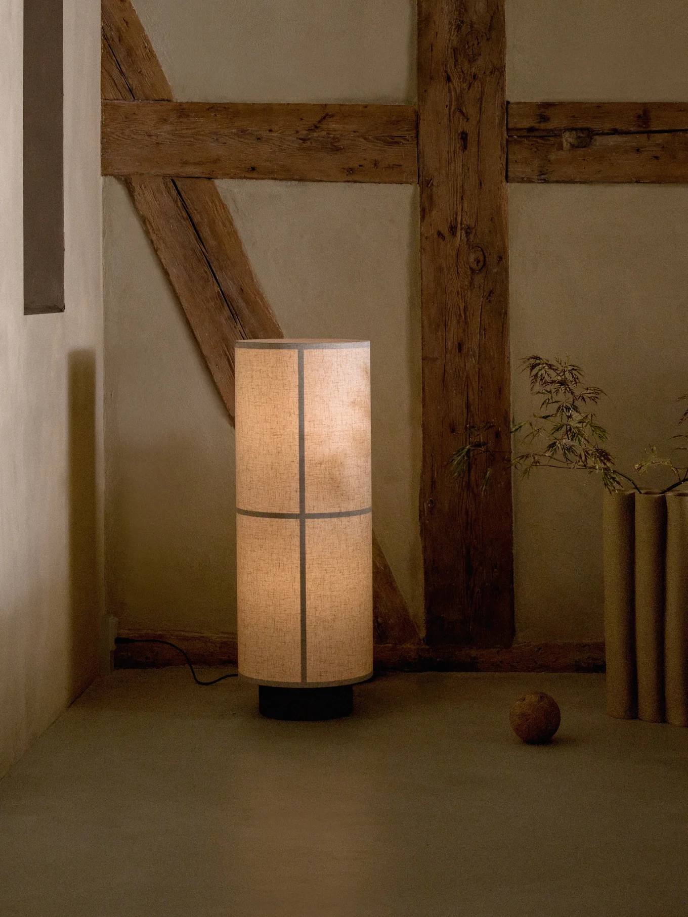 Hoge vloerlamp met gesegmenteerde stoffen kap, die een warme gloed werpt in een rustieke kamer met zichtbare houten balken.
