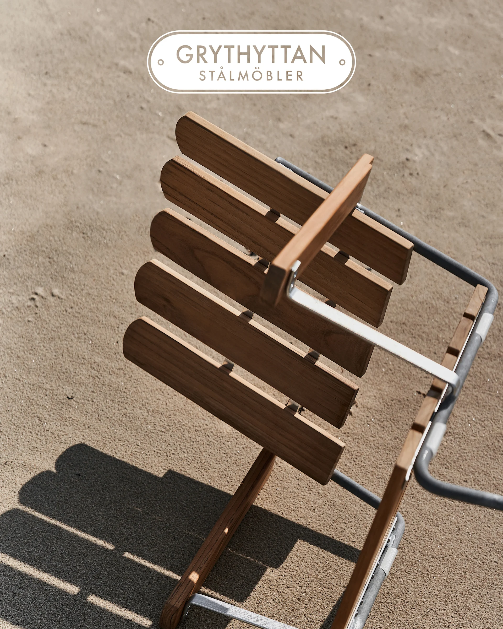Houten lattenstoel met metalen frame op zand, van bovenaf gezien. Grythyttan Stålmöbler logo.