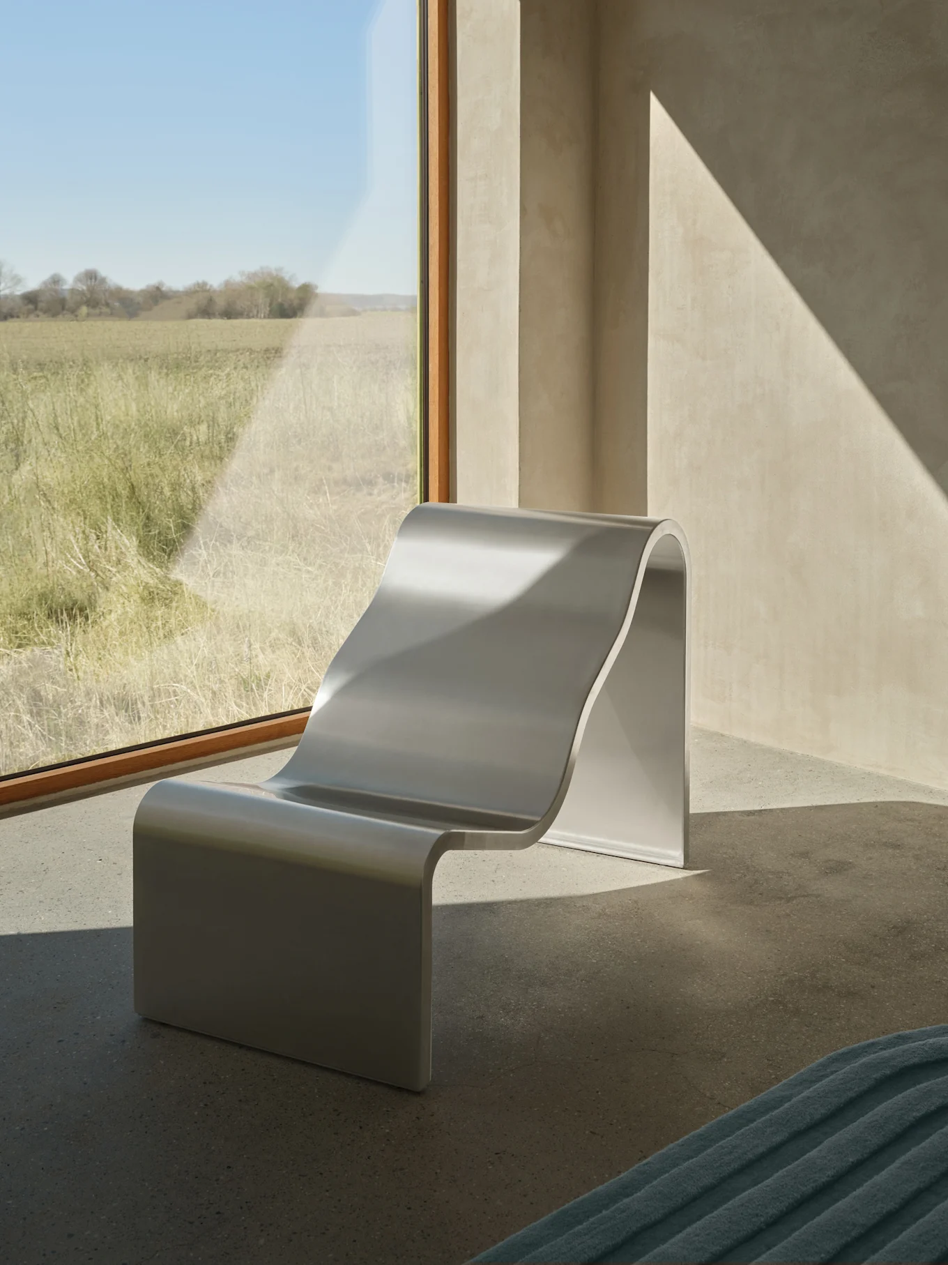 Een moderne zilverkleurige S-vormige loungestoel staat bij een groot raam met uitzicht op een zonnig grasveld, met diagonale zonlichtpatronen op de muren.