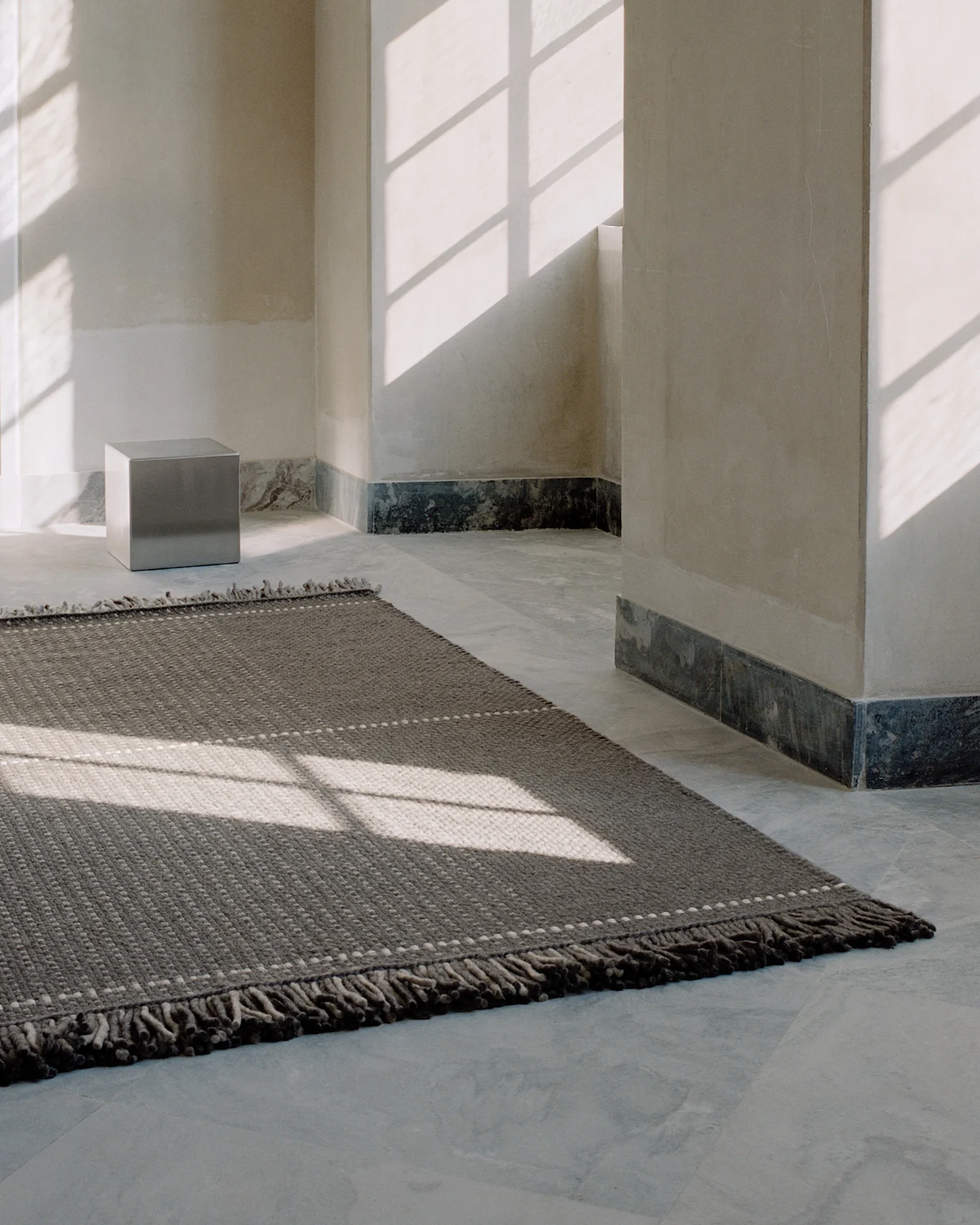 Een donkergrijs getextureerd vloerkleed met lichte stippen op een lichte tegelvloer, verlicht door raamschaduwen in een minimalistische kamer.