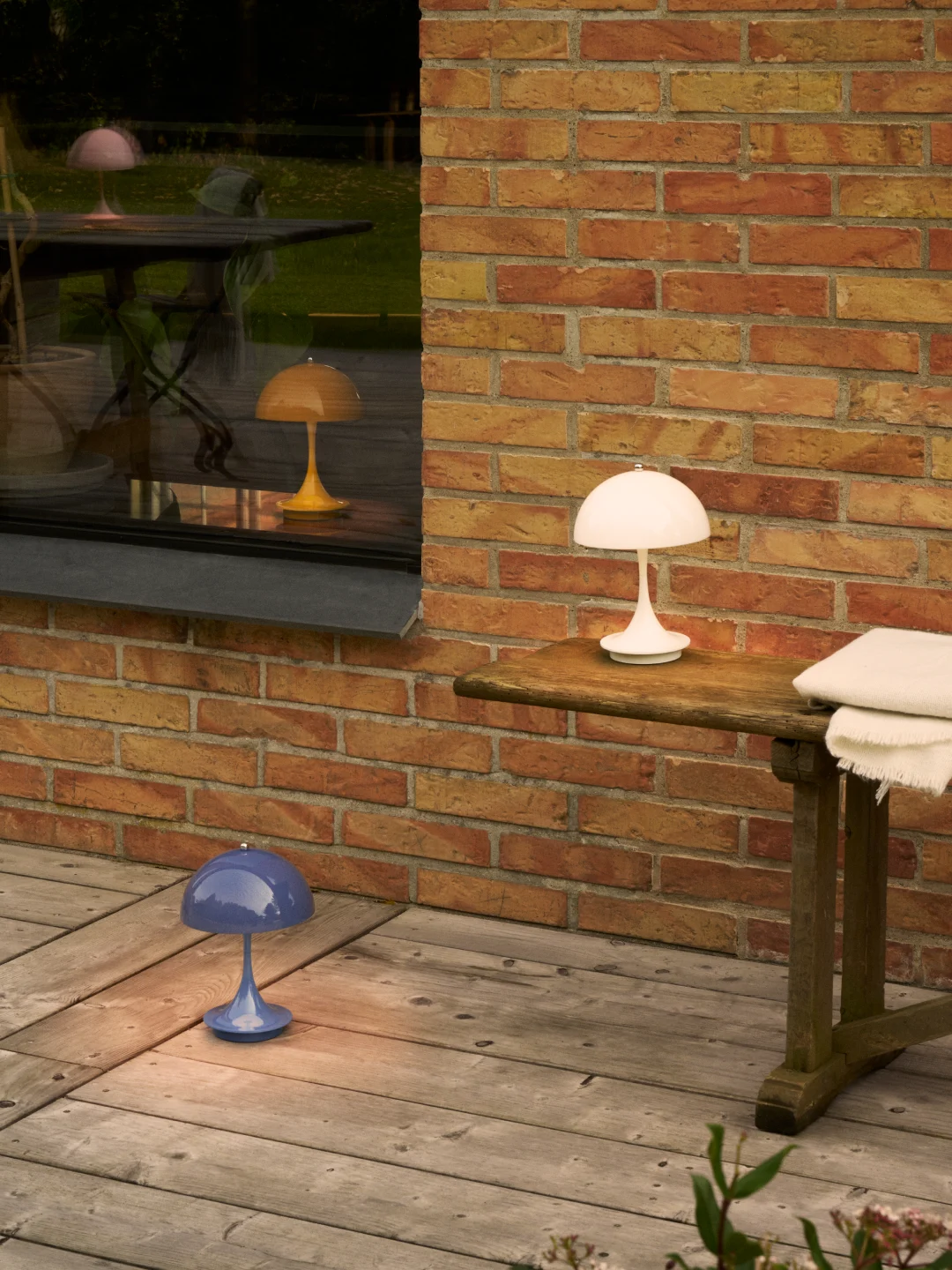 Witte paddenstoellamp op tafel, blauwe op vlonder. Roze en gele paddenstoellamp reflecties in raam bij bakstenen muur.