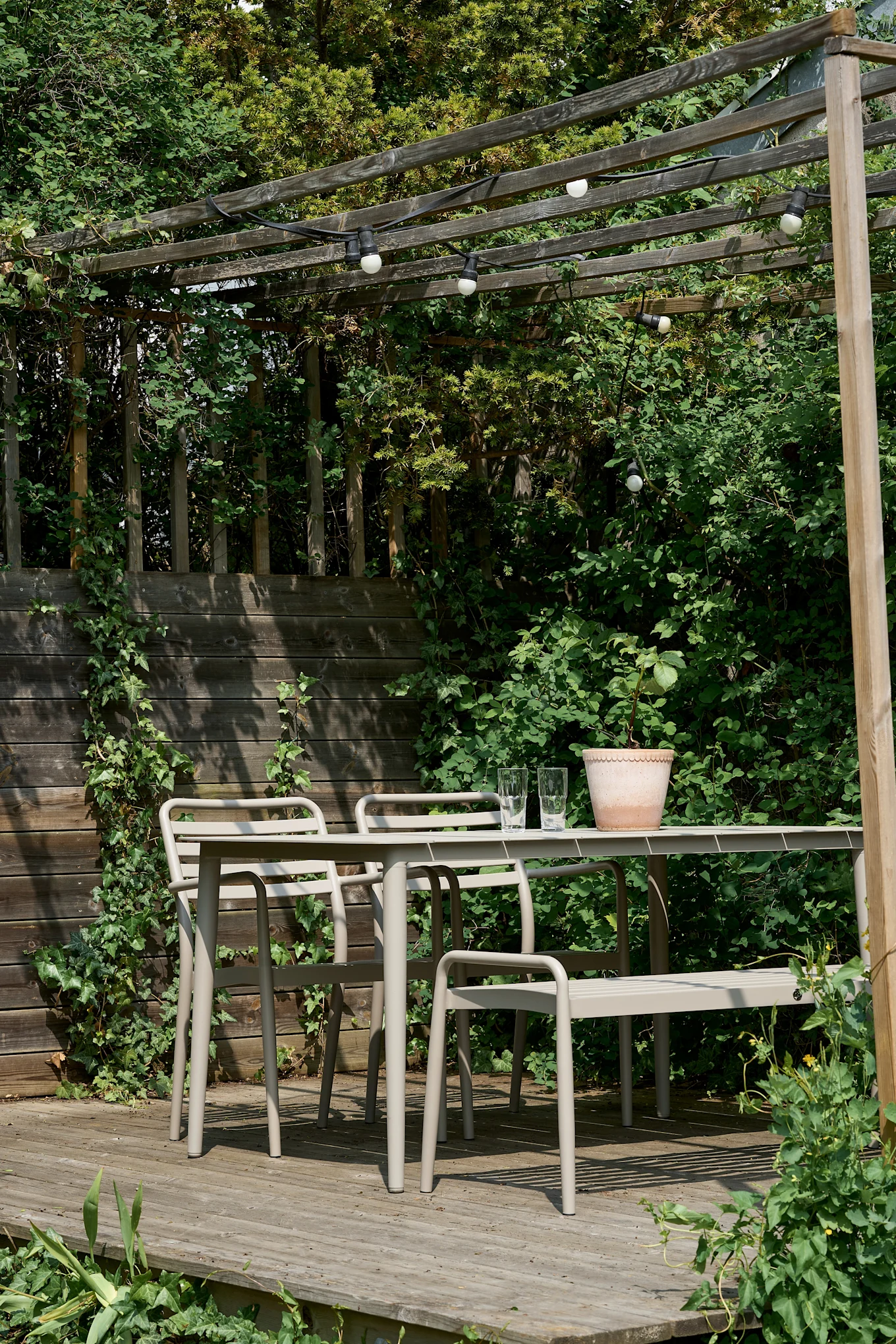 Een buiten eethoek met een beige metalen tafel en stoelen op een houten terras onder een pergola met lichtslingers.