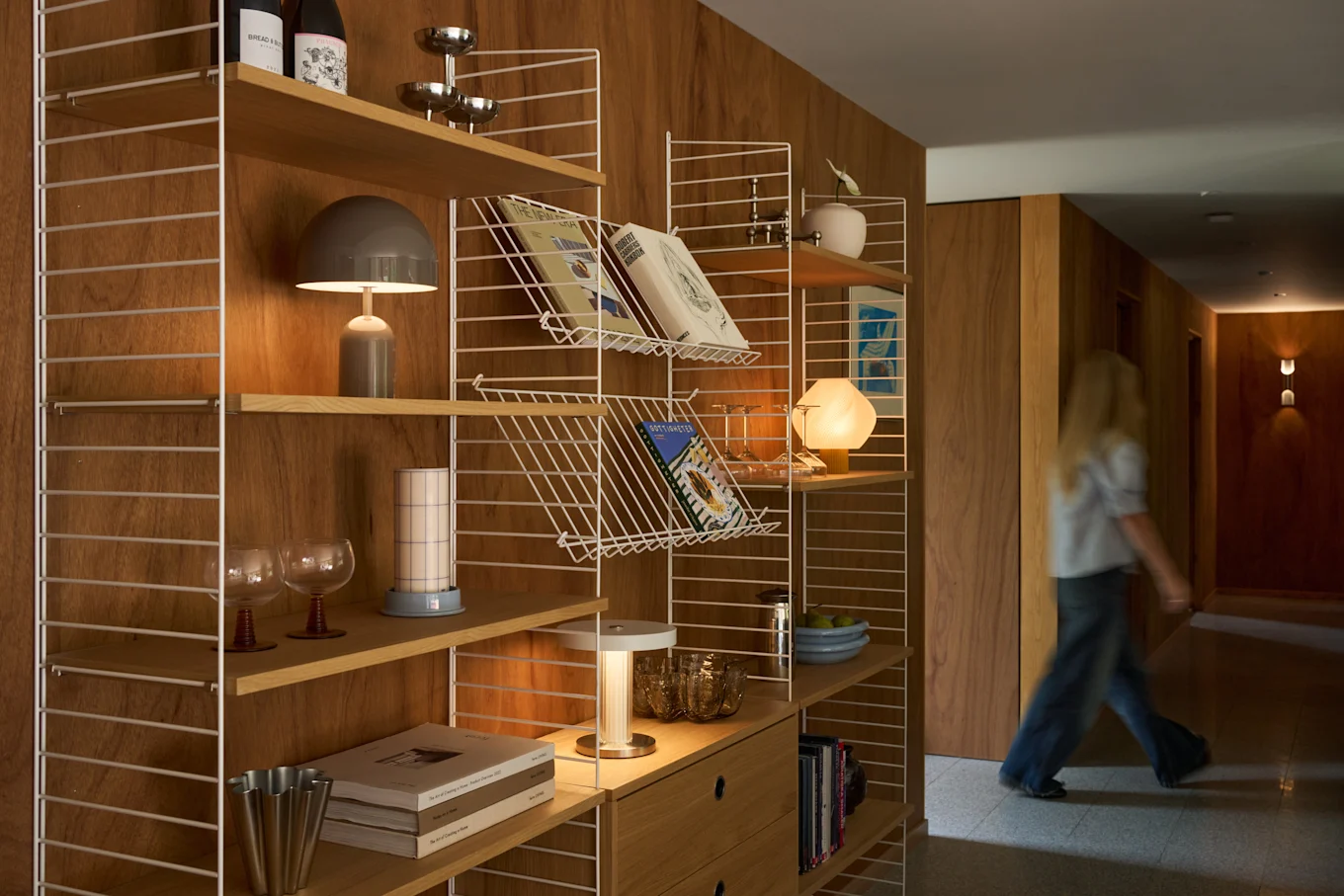Een blond persoon loopt door een houten gang naast een moderne wandkast gevuld met boeken, lampen en decoratieve items.