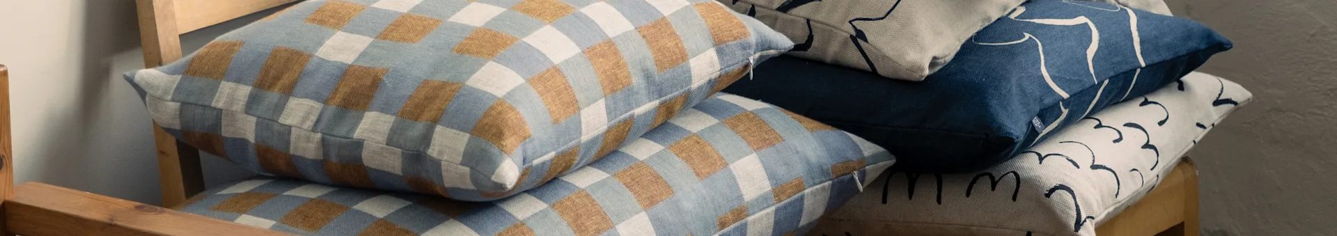 Gestapelde kussens op een houten stoel, met geruite blauwe en bruine, en abstracte blauwe en beige patronen.