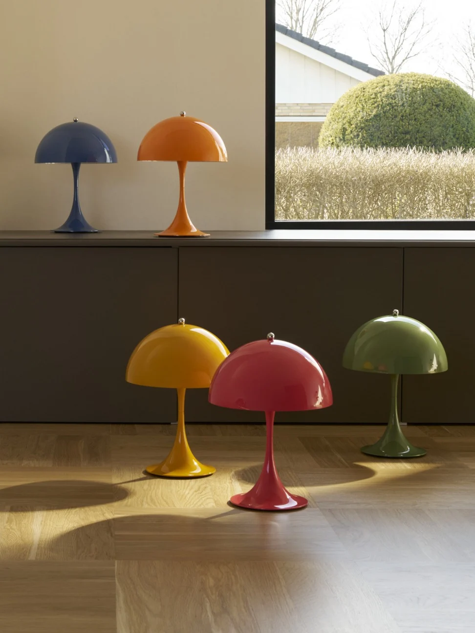 Zes kleurrijke paddestoelvormige lampen, twee op een kast en vier op een houten vloer, bij een raam dat uitkijkt op een tuin.