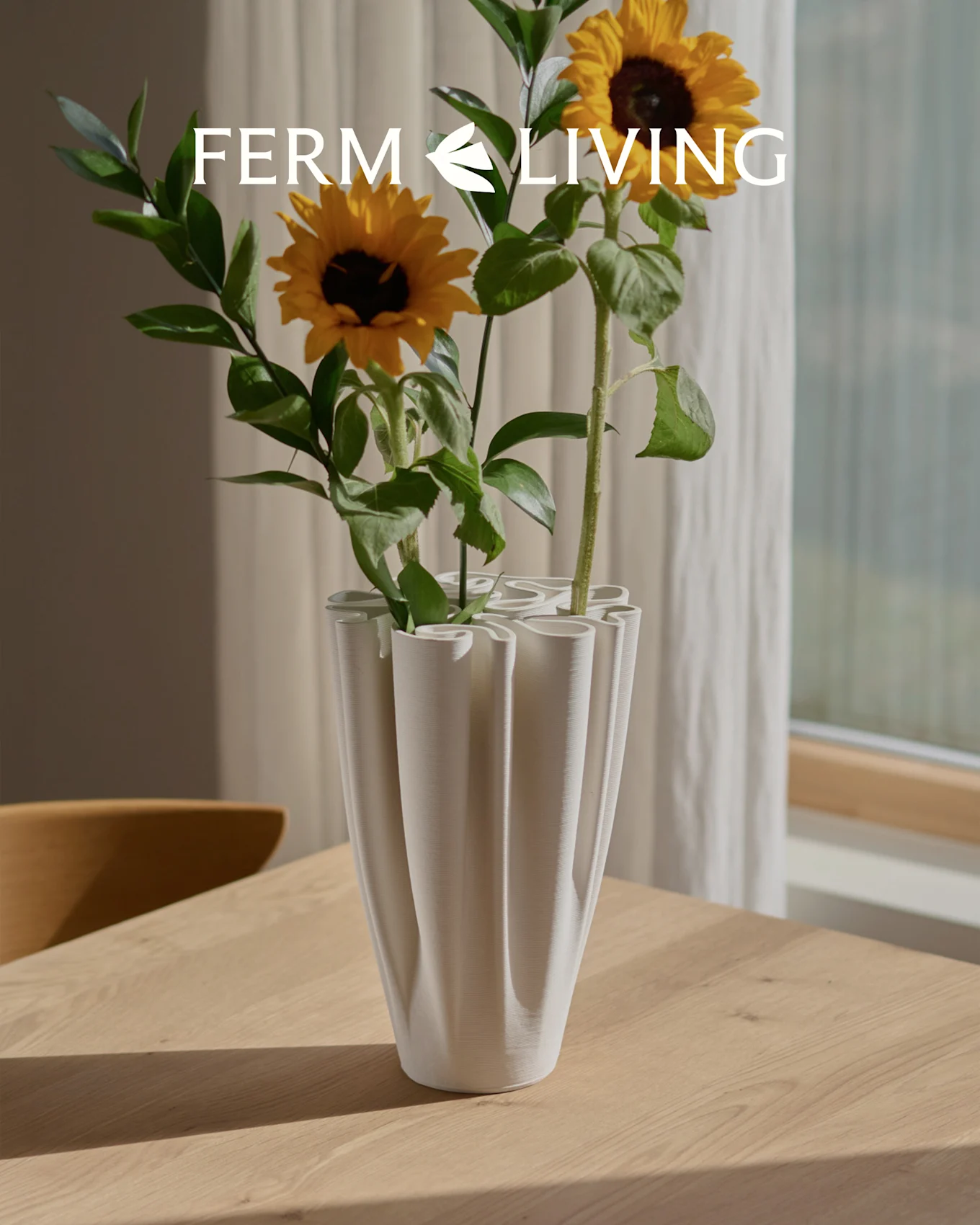 Zonnebloemen in een witte, geribbelde golvende vaas op een houten tafel. Ferm Living logo overlay.