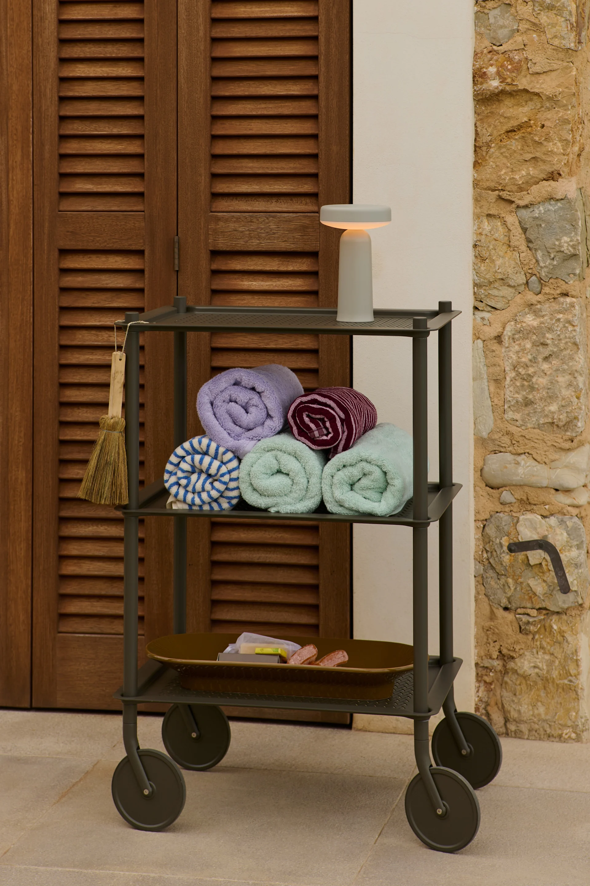 Een drielaagse trolley met opgerolde handdoeken, toiletartikelen en een lamp naast een houten deur en stenen muur.