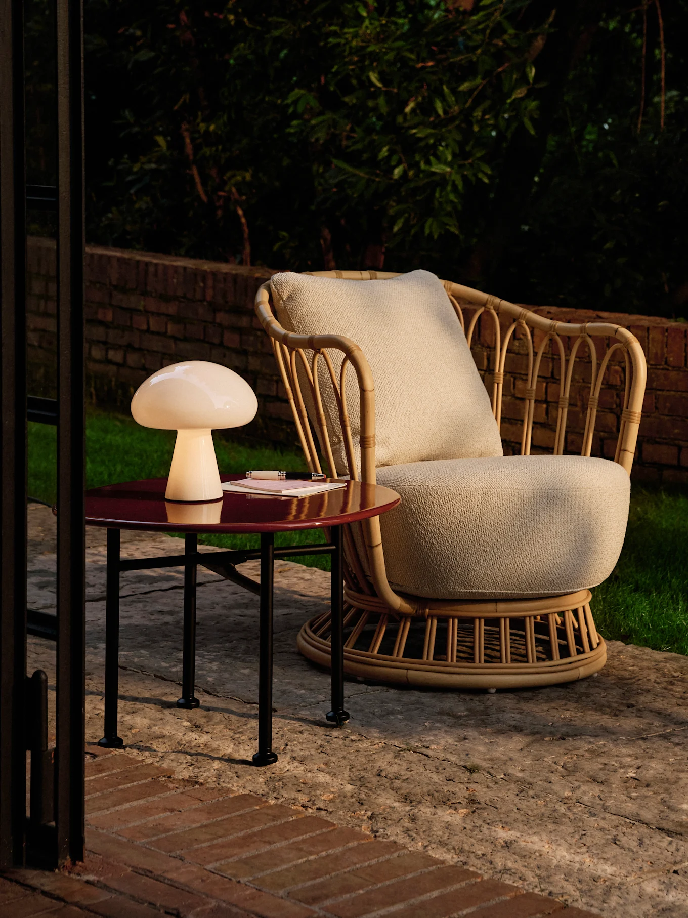 Paddestoellamp op een rode bijzettafel naast een rotan fauteuil op een terras in de schemering.