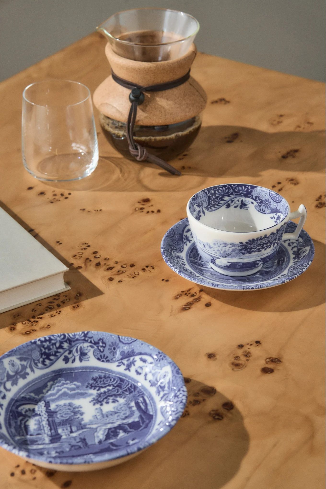 Een pour-over koffiezetapparaat, een leeg glas, een blauw-wit theekopje op een schotel, en een bijpassend bord op een lichte houten tafel.