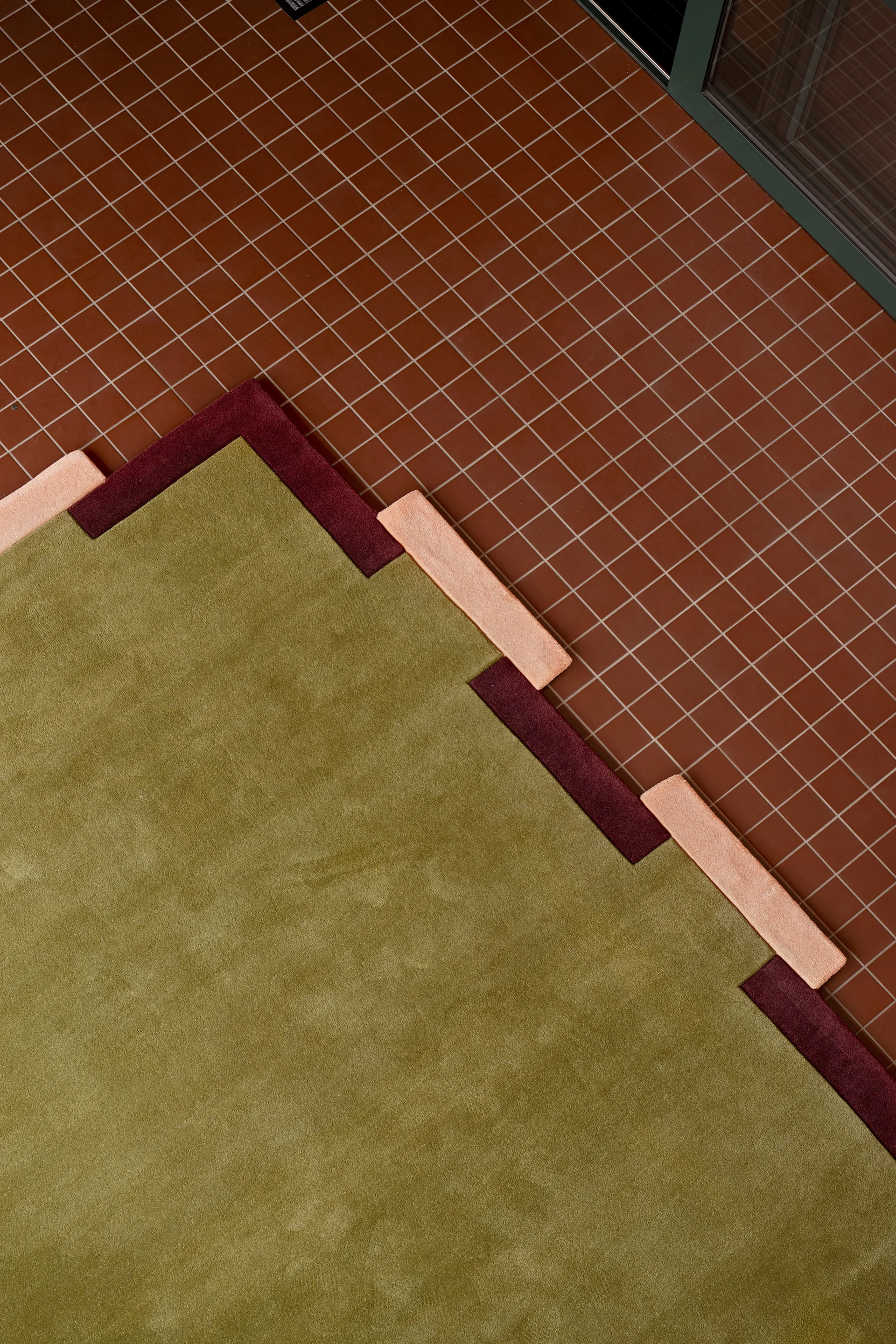 Een geometrisch vloerkleed in groen, bordeauxrood en perzik met een getrapt patroon, liggend op een roodbruine tegelvloer.