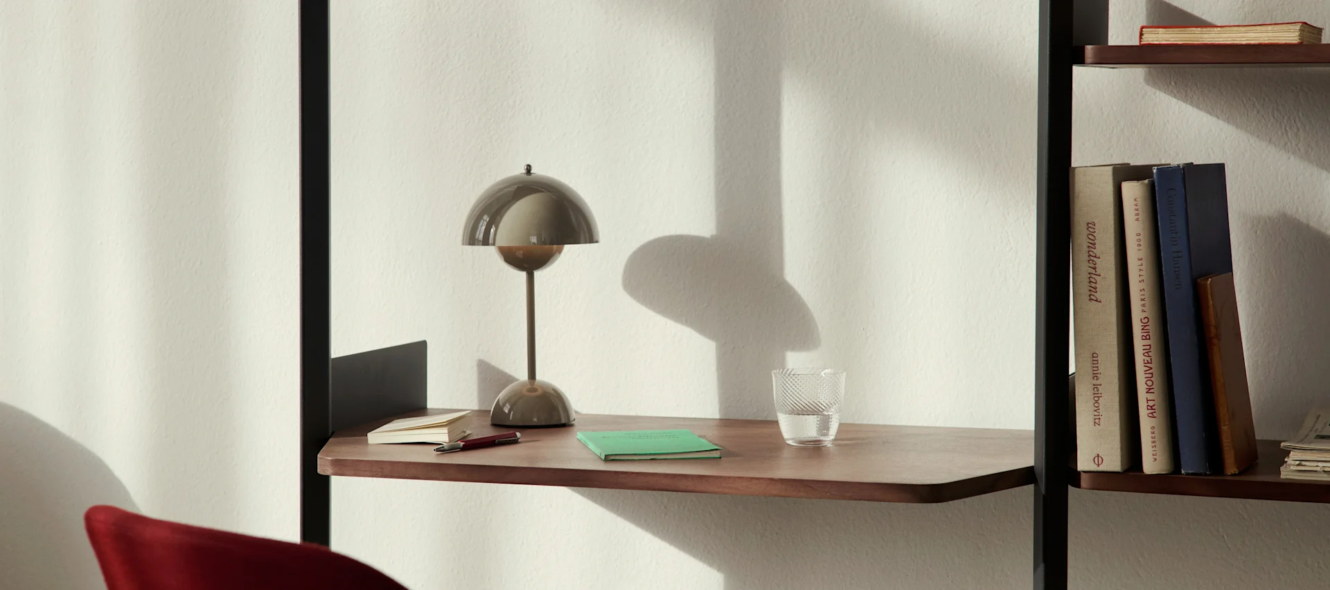 Een moderne paddestoelvormige tafellamp, een glas water en notitieboekjes op een houten bureauschap naast een boekenkast.