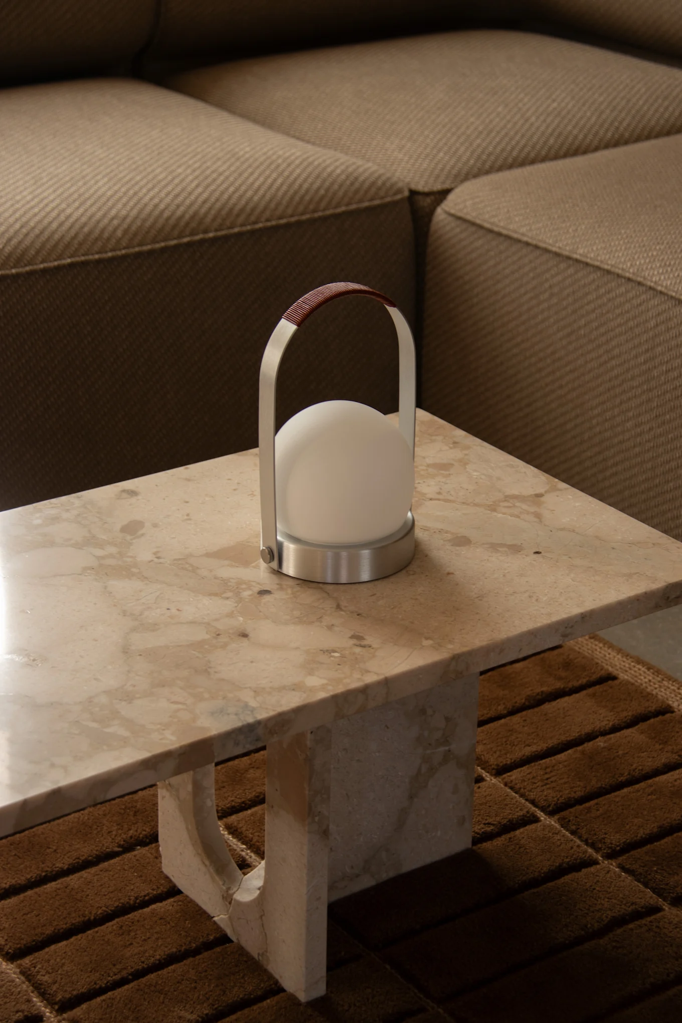 Moderne draagbare lamp met wit bolvormig licht, geborsteld metalen frame en bruin lederen handvat op lichte marmeren tafel.