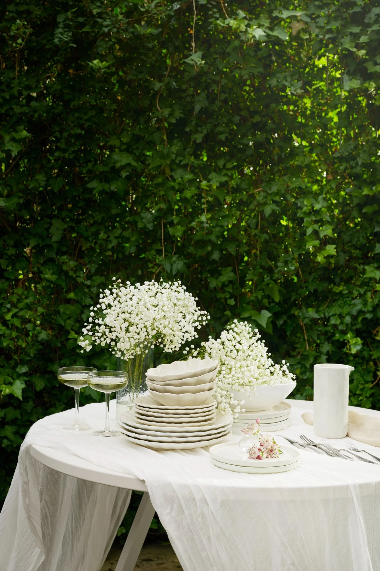 Buitentafel gedekt met wit servies, gipskruidbloemen, champagneglazen en bestek op een wit tafelkleed, tegen weelderig groene bladeren.