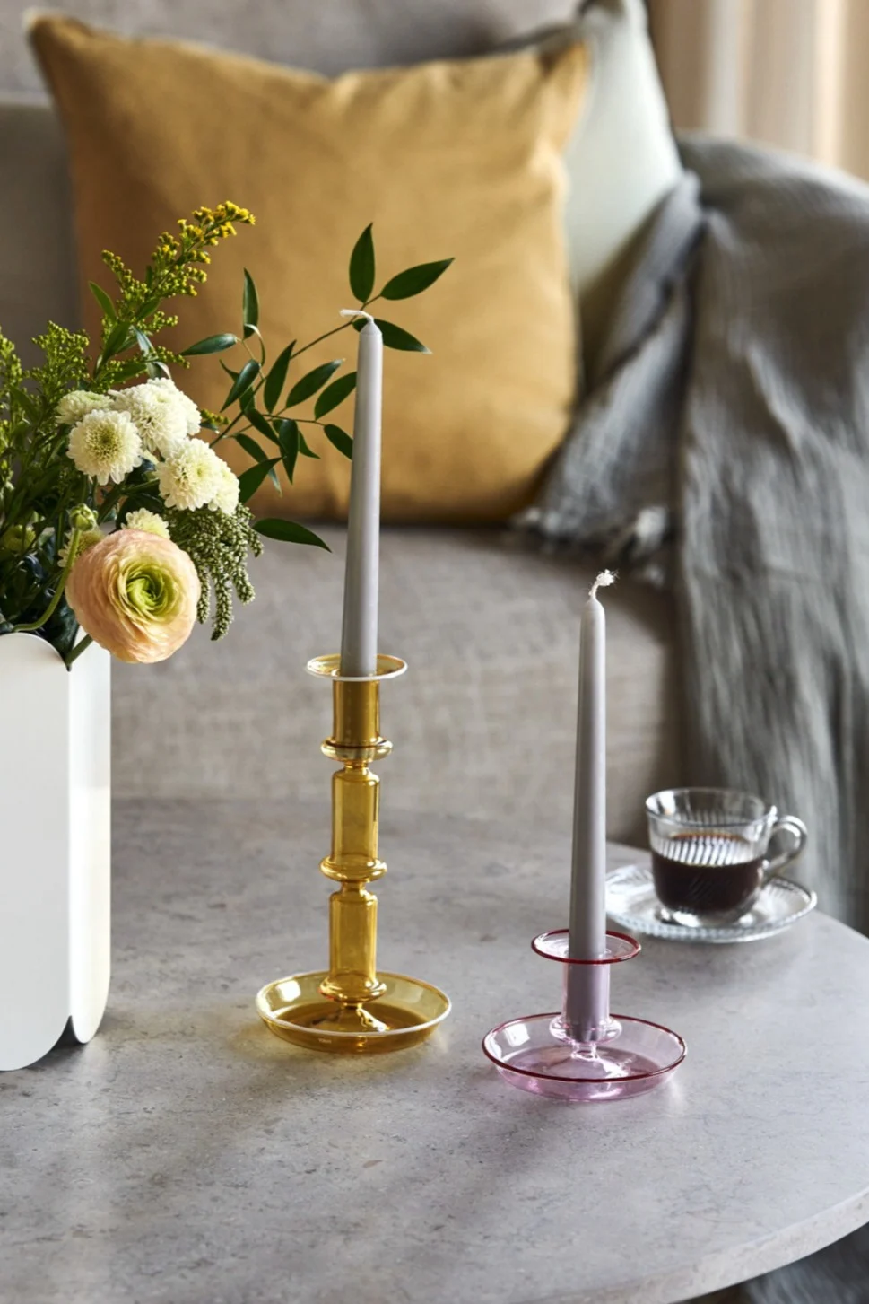 Twee glazen kandelaars (amber en roze) met grijze kaarsen op een tafel, naast een vaas bloemen en een kop koffie.
