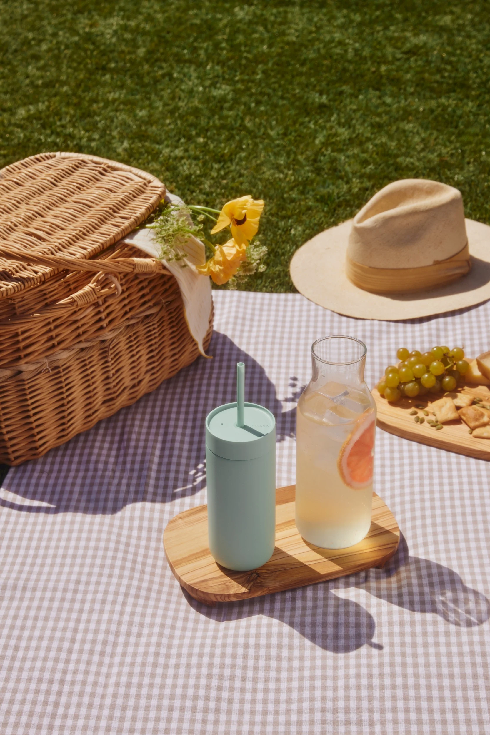 Picknick op een geruite deken: rieten mand, lichtgroene beker, grapefuitdrankje, druiven en crackers op het gras.