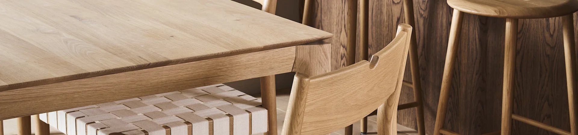 Close-up van een lichte houten eettafel met een stoel met gevlochten zitting en houten krukken op de achtergrond.