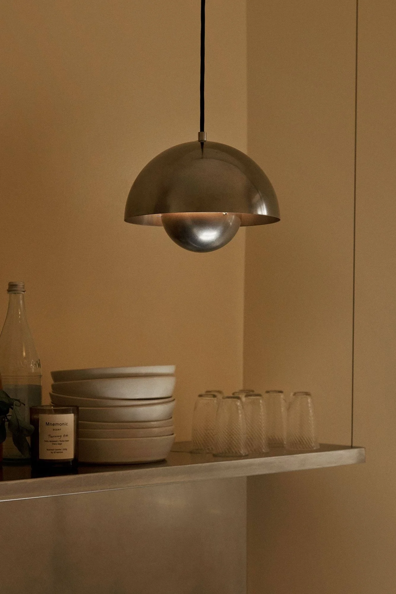 Metalen koepelvormige hanglamp verlicht plank met gestapelde kommen, glazen, een fles en een kaars in warme kamer.