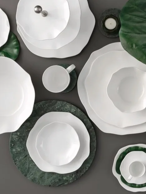 Een collectie wit keramisch servies met golvende randen, gerangschikt met groene bladeren en marmeren onderzetters op een donker oppervlak.