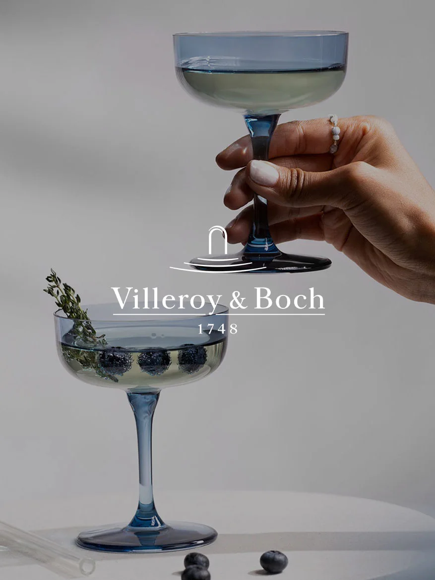 Hand houdt blauw coupeglas vast; een ander glas eronder met bosbessen, tijm en Villeroy & Boch logo en '1748'.