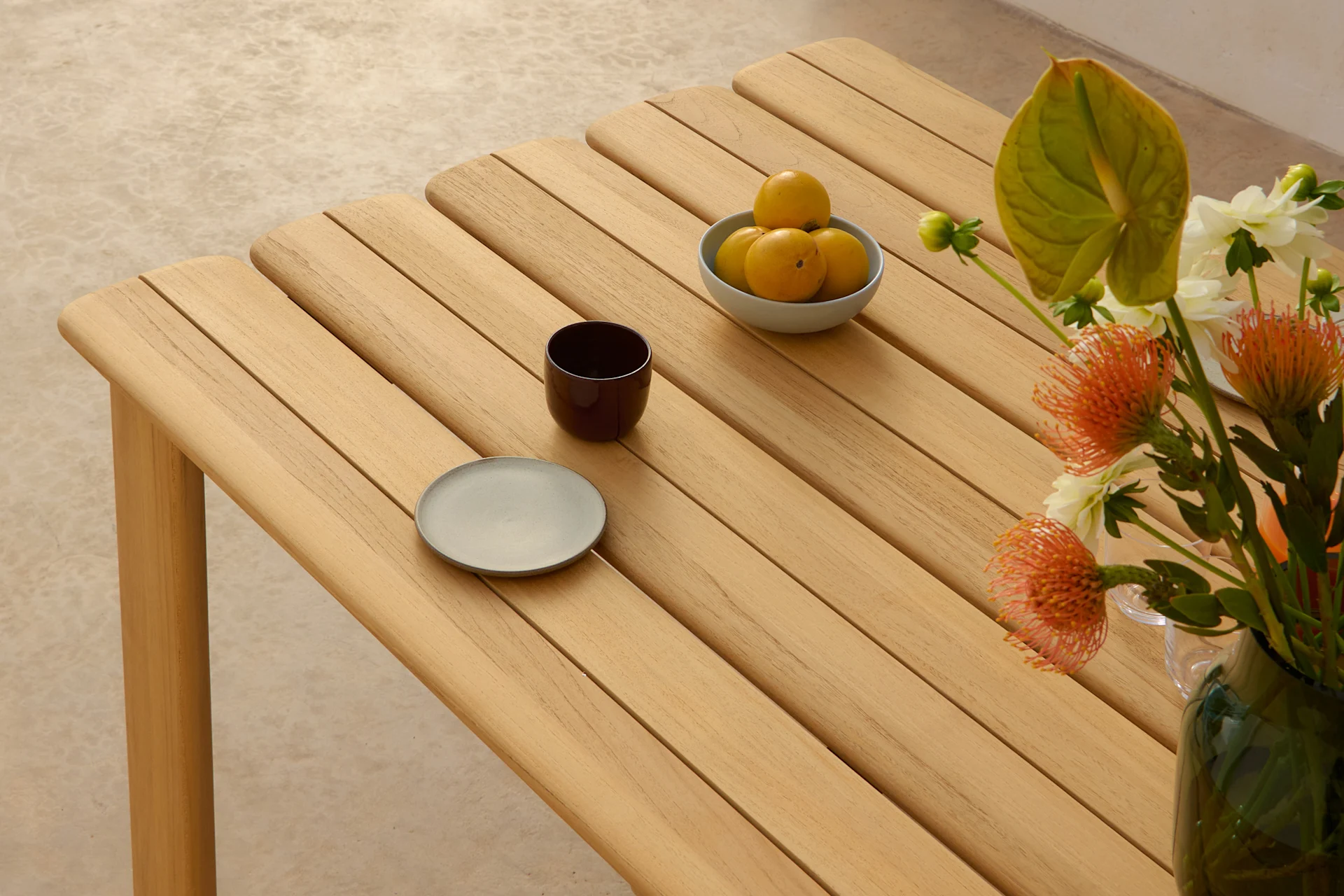 Een lichte houten lamellentafel met een kom gele vruchten, een donkere kop, een grijs bord en een vaas met oranje en witte bloemen.