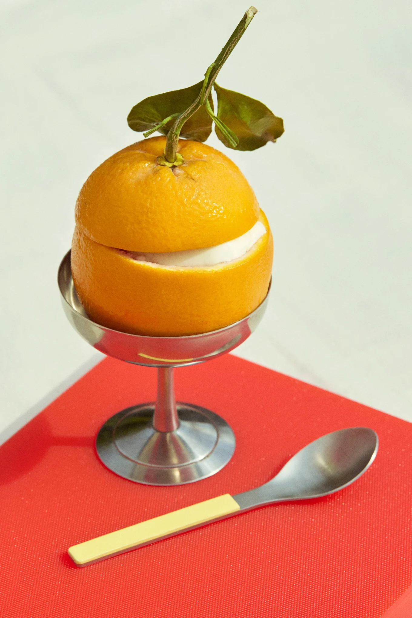 Sinaasappel dessert met witte vulling in zilveren beker, met lepel, op rood oppervlak.