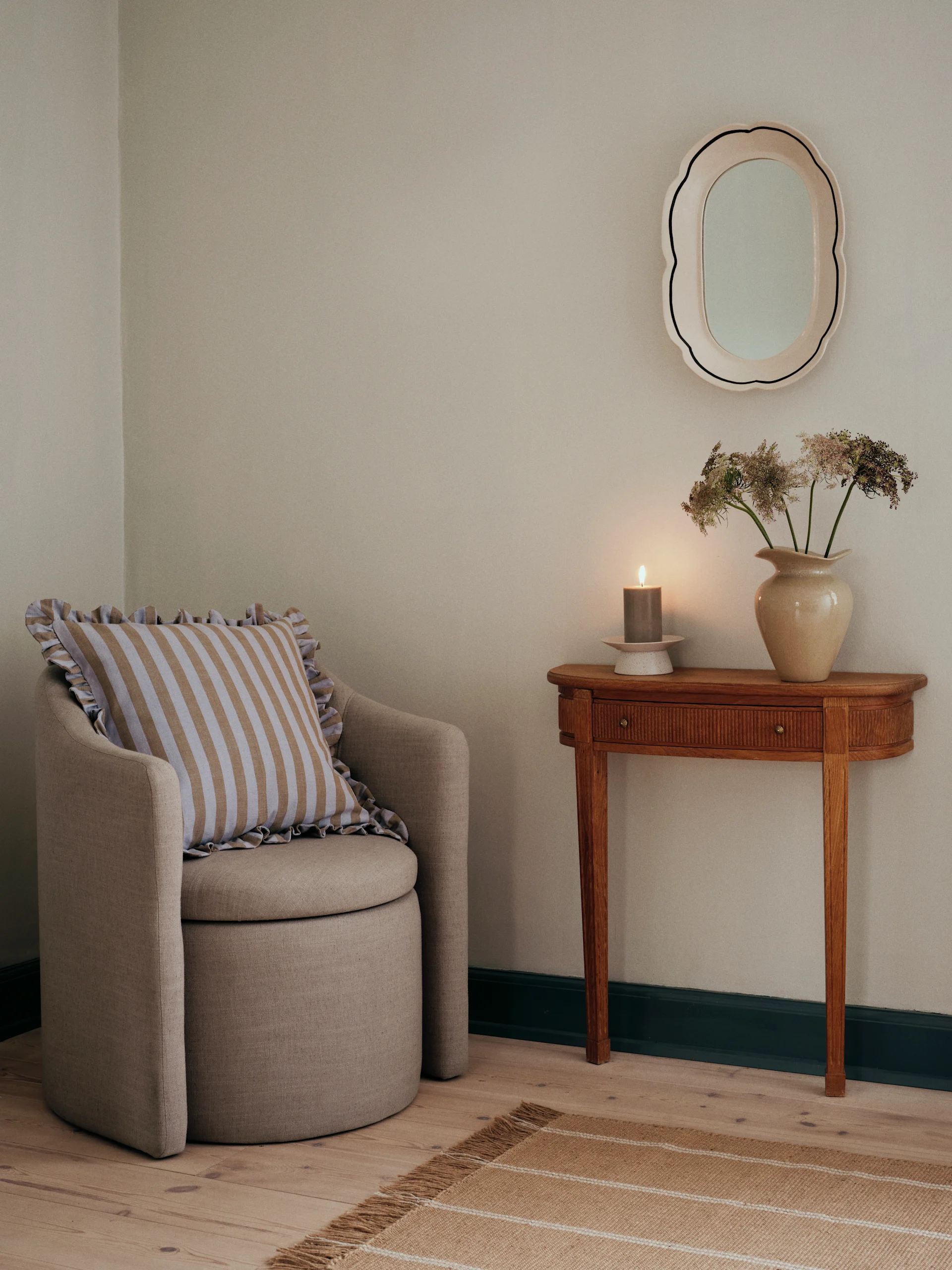 Een fauteuil met een gestreept kussen naast een houten sidetable met een brandende kaars, gedroogde bloemen en een geschulpte spiegel.