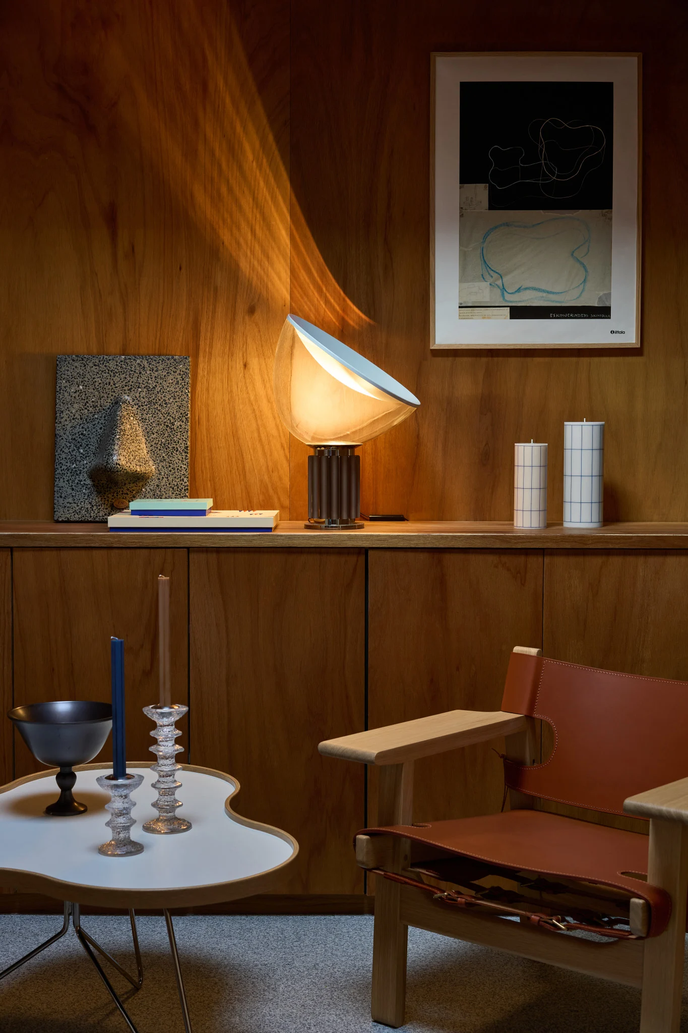 Een lamp verlicht een houten kamer met planken, kunst, kaarsen, een witte tafel en een leren fauteuil.