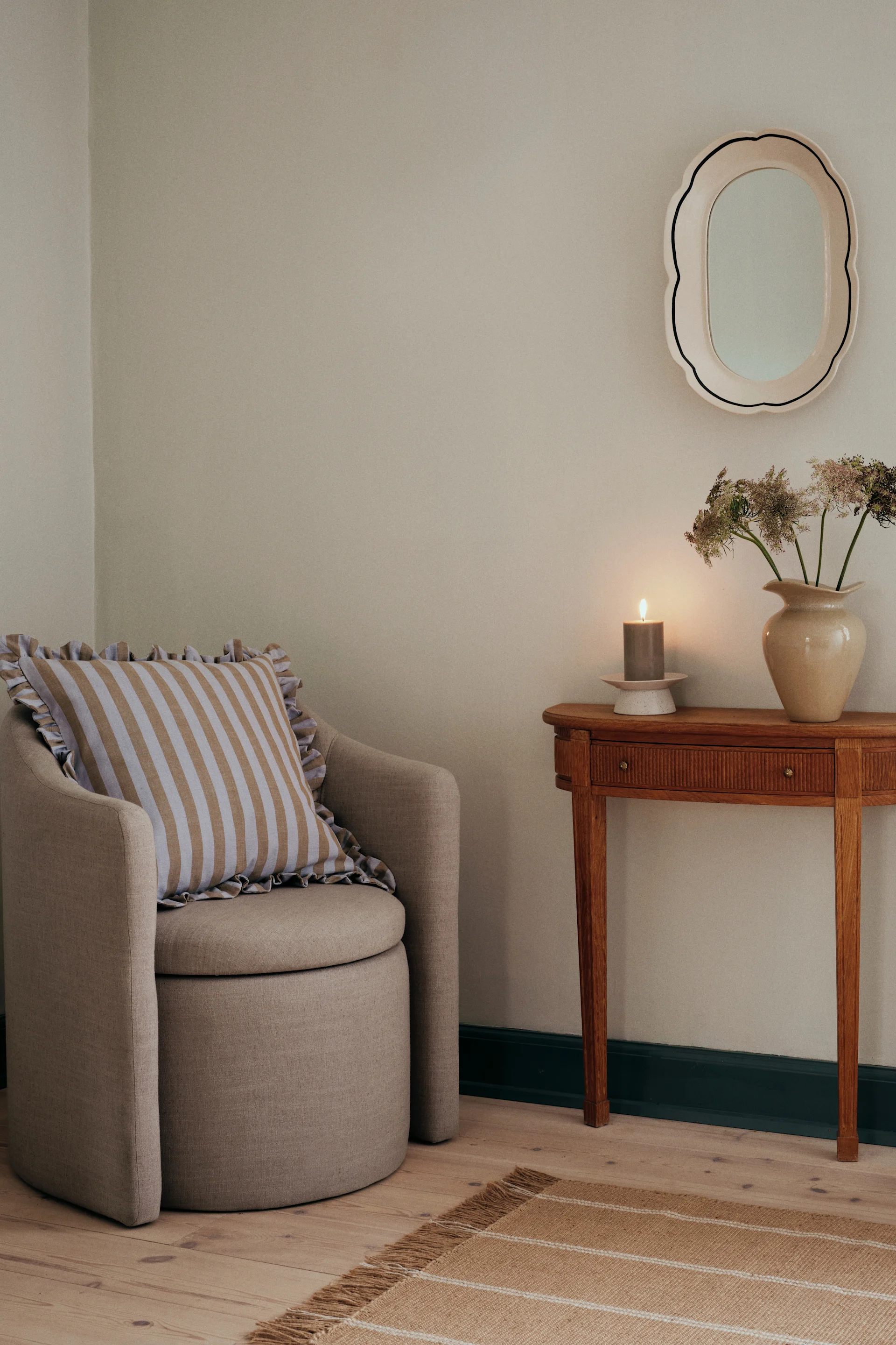 Een fauteuil met gestreept kussen, een houten bijzettafel met brandende kaars en vaas bloemen, een spiegel en een gestreept vloerkleed.