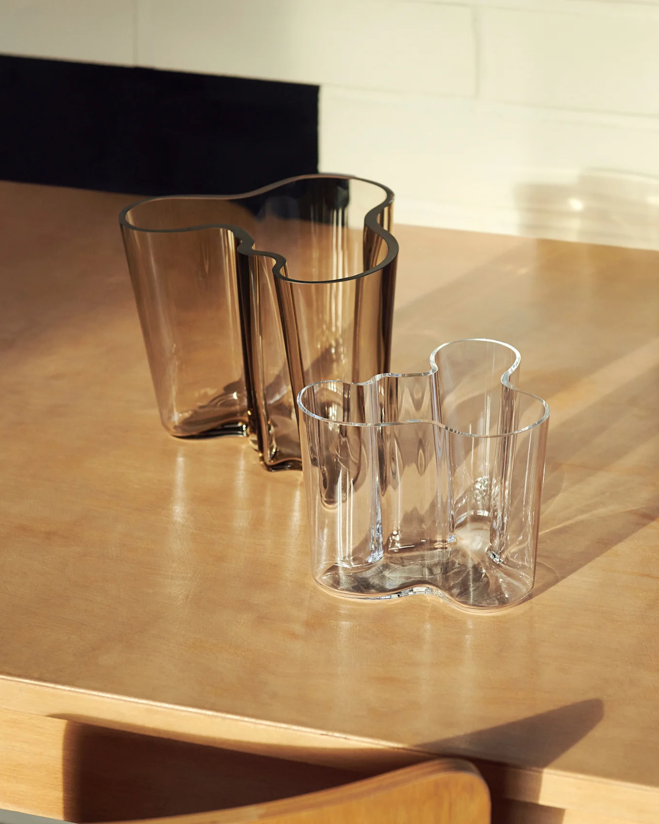 Twee golvende glazen vazen, één grote amberkleurige en één kleinere heldere, op een houten oppervlak.