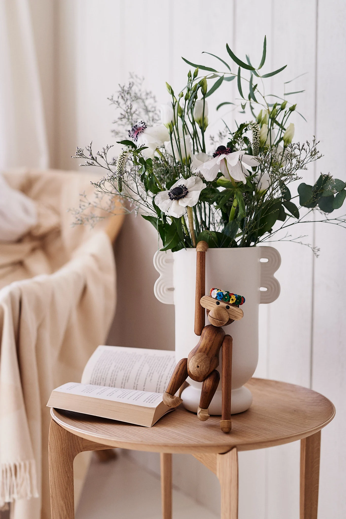 Een houten aapspeeltje hangt aan een witte vaas met witte bloemen naast een open boek op een houten tafel.