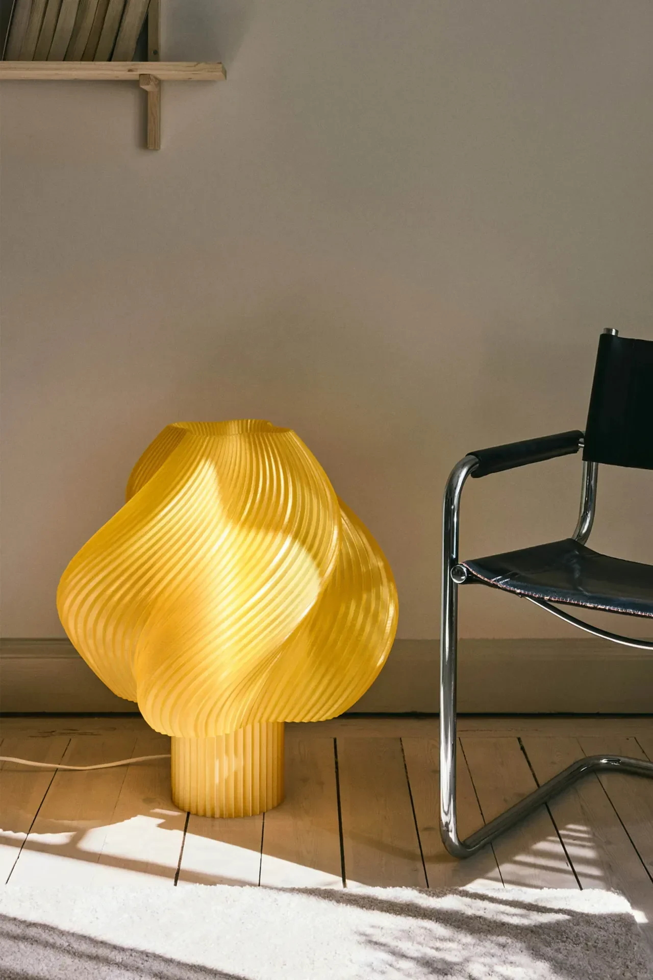 Een felgele, sculpturale tafellamp met een geribbeld, golvend ontwerp schijnt op een houten vloer naast een zwartleren en chromen stoel.