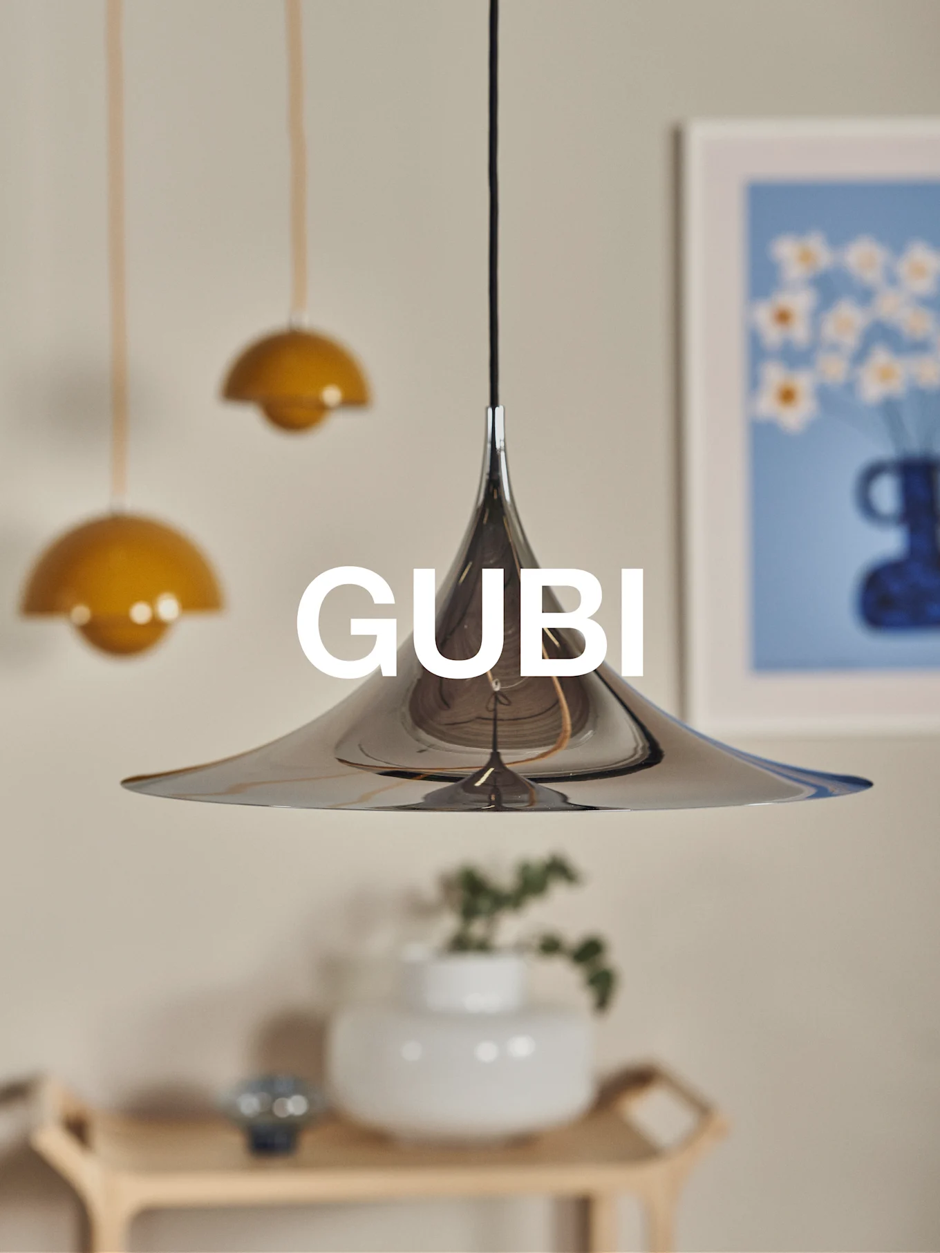 Een gepolijste chromen conische hanglamp met het GUBI-logo, tegen een wazige achtergrond met andere lampen en kunst.