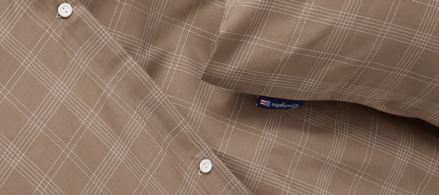 Close-up van een bruine stof met wit ruitpatroon, met twee witte knopen en een blauw Lexington-label.