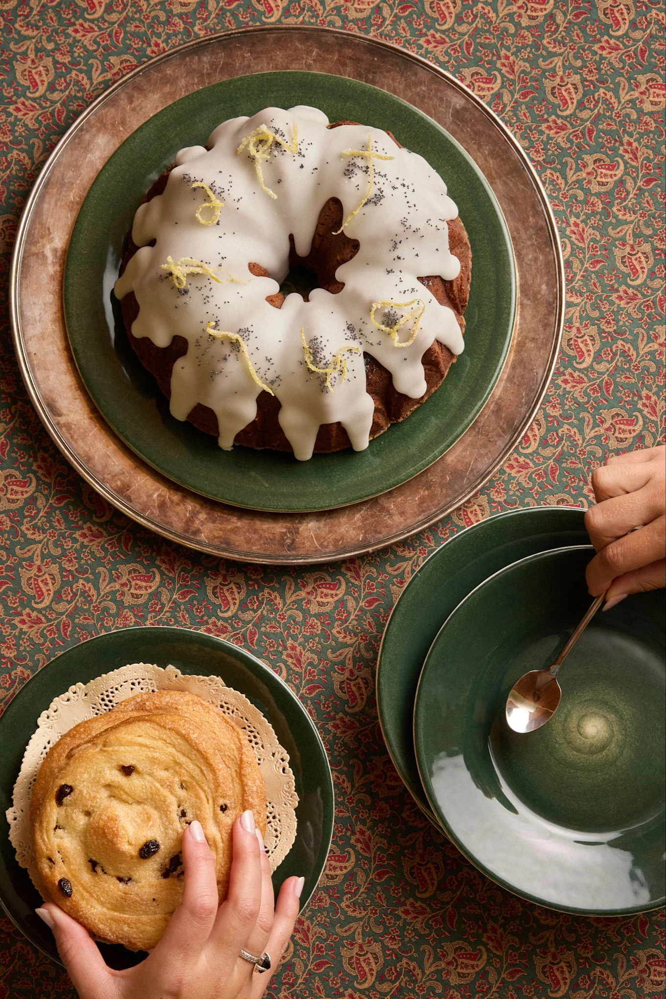 Bovenaanzicht: Bundtcake met citroen en maanzaad, rozijnenbroodje en handen op een gedessineerd tafelkleed.