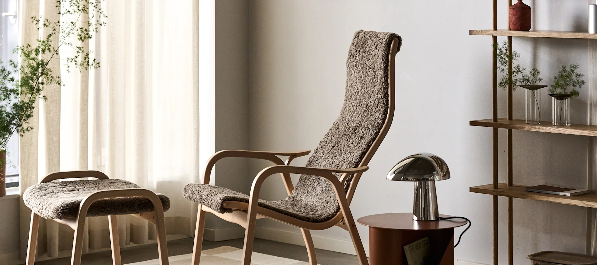 Een lichte houten fauteuil en kruk met getextureerde bruine bekleding, een metalen tafellamp en een houten plank met planten.