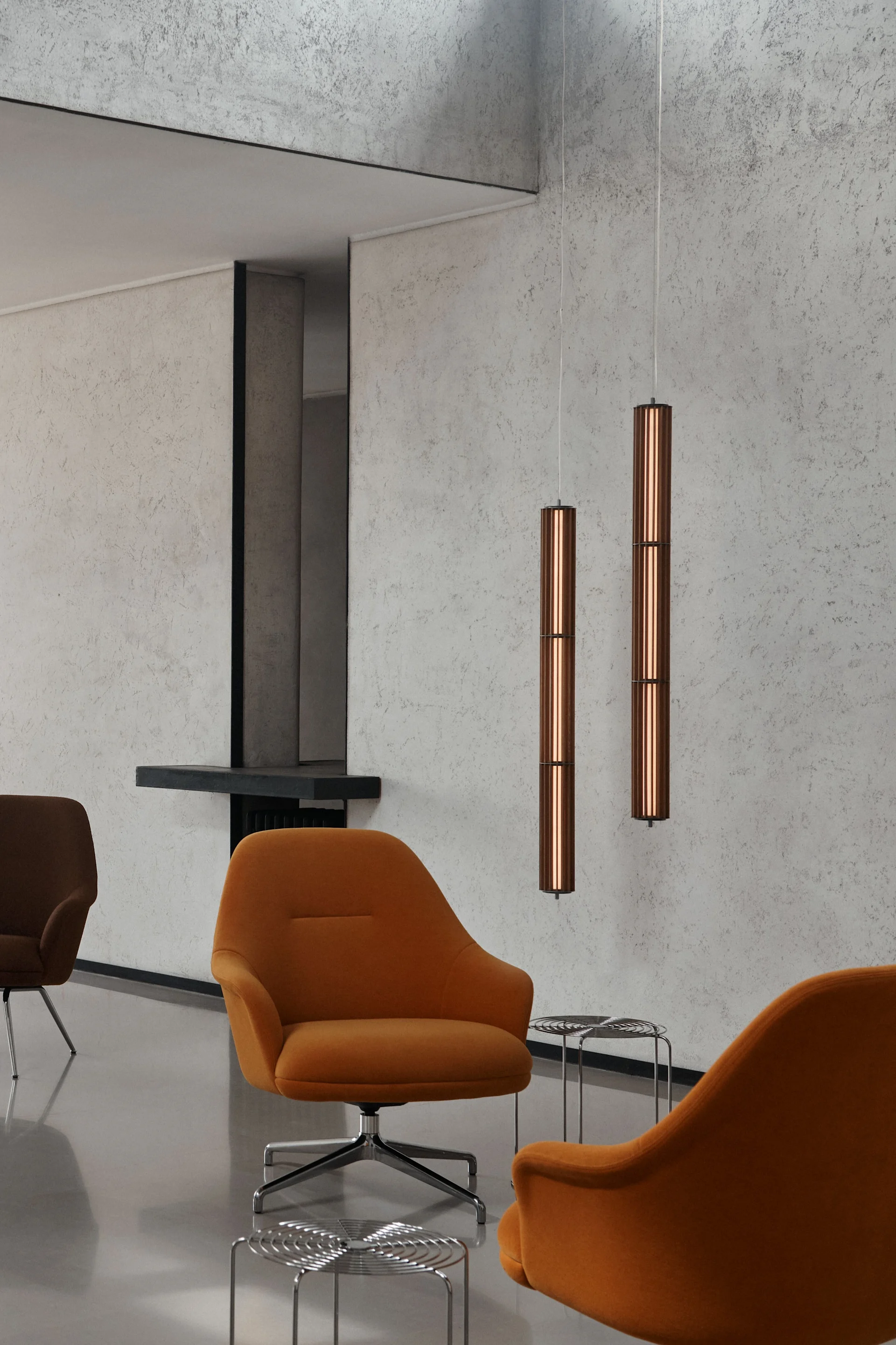 Twee cilindrische koperen hanglampen in een moderne kamer met oranje fauteuils en wanden met betontextuur.