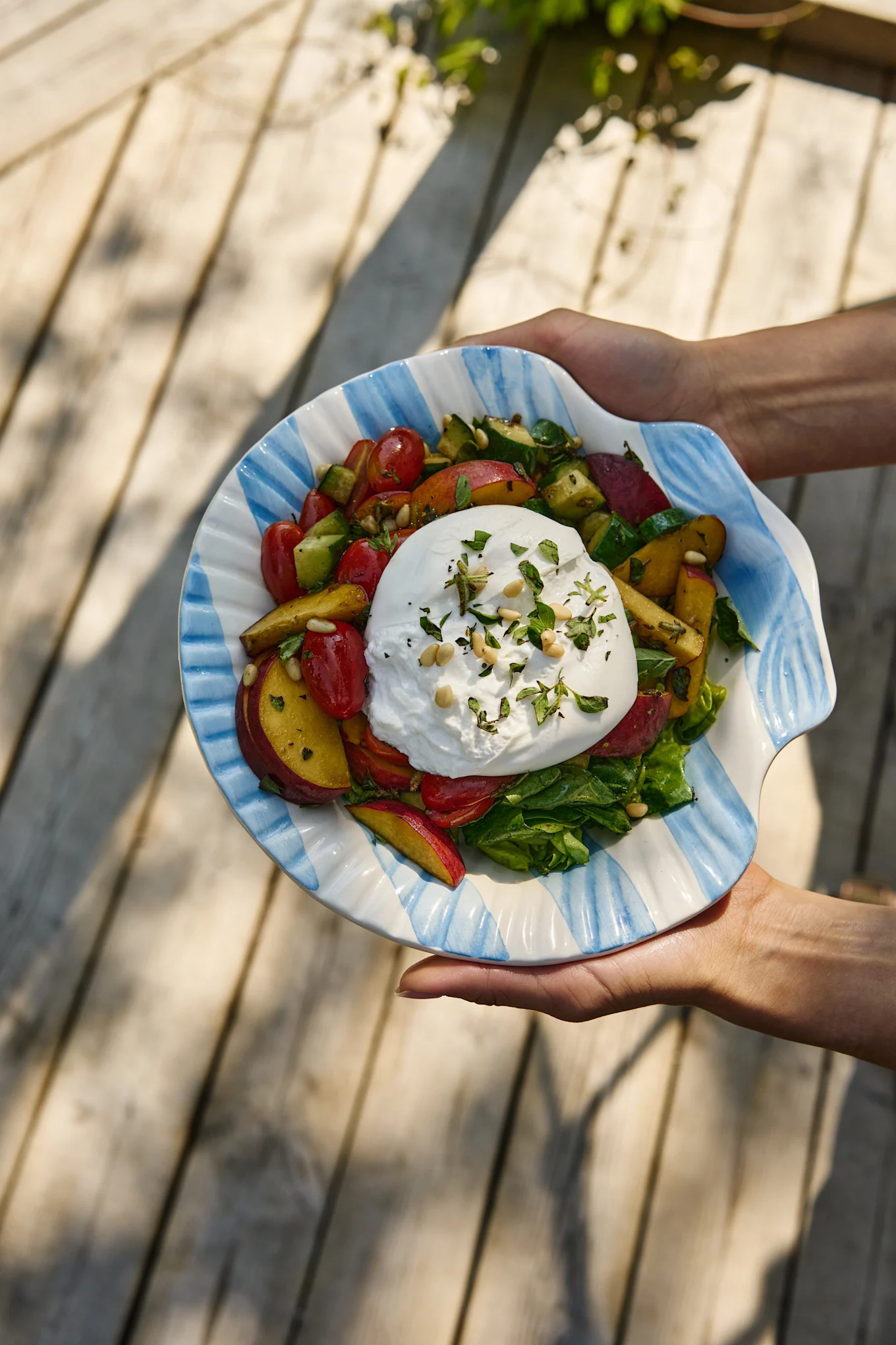Handen houden een burrata salade met tomaten, perziken, komkommer en groenten in een gestreepte schelpvormige kom.