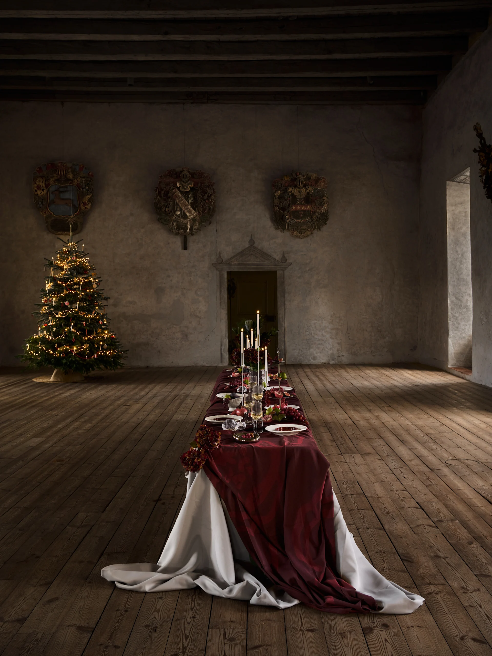 Lange kerstdinertafel met rood tafelkleed, kaarsen, fruit en servies, naast een verlichte kerstboom in een oude hal.