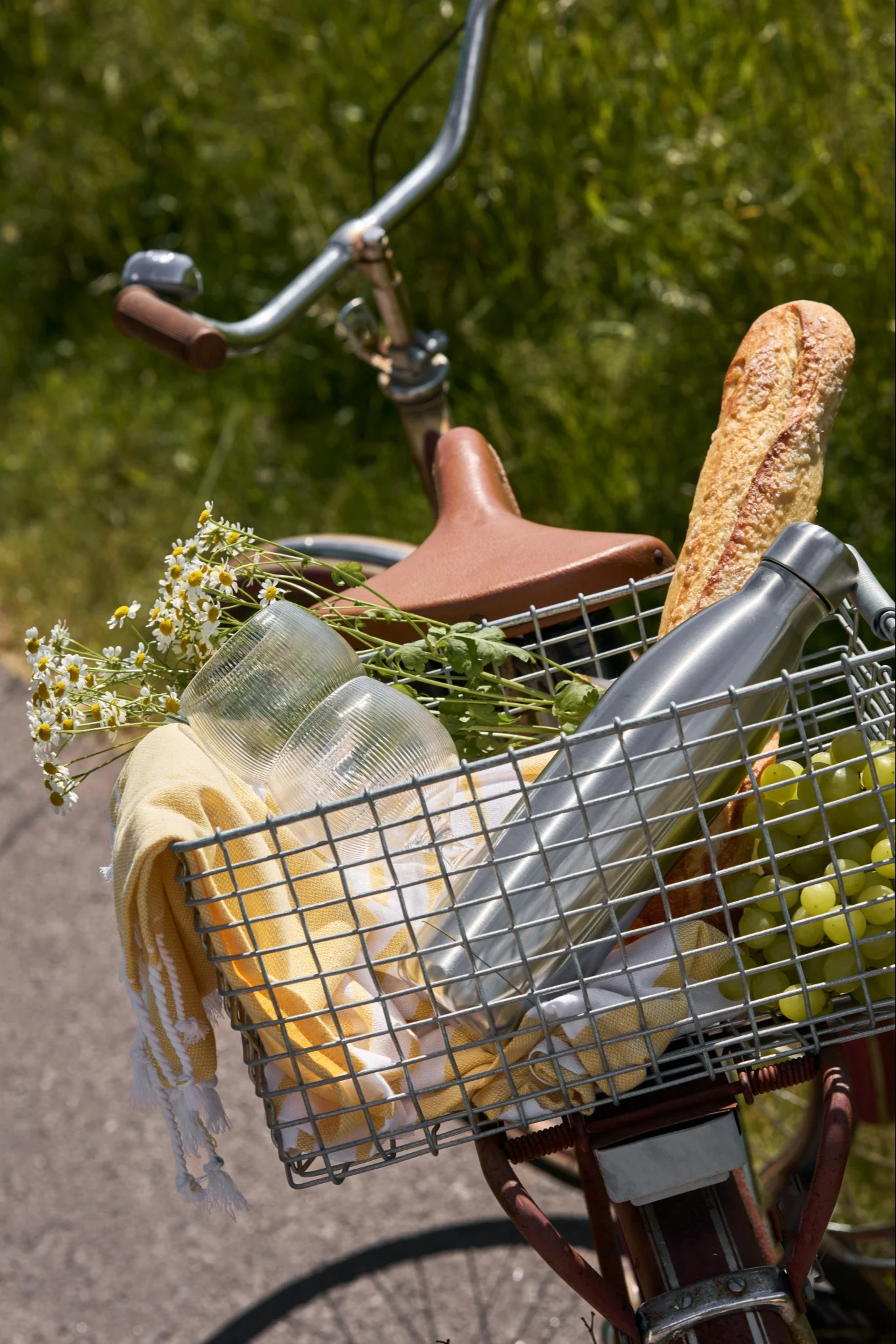 Fiets met een voormand gevuld met picknickspullen: brood, groene druiven, een zilveren waterfles, bekers, een gele handdoek en bloemen.