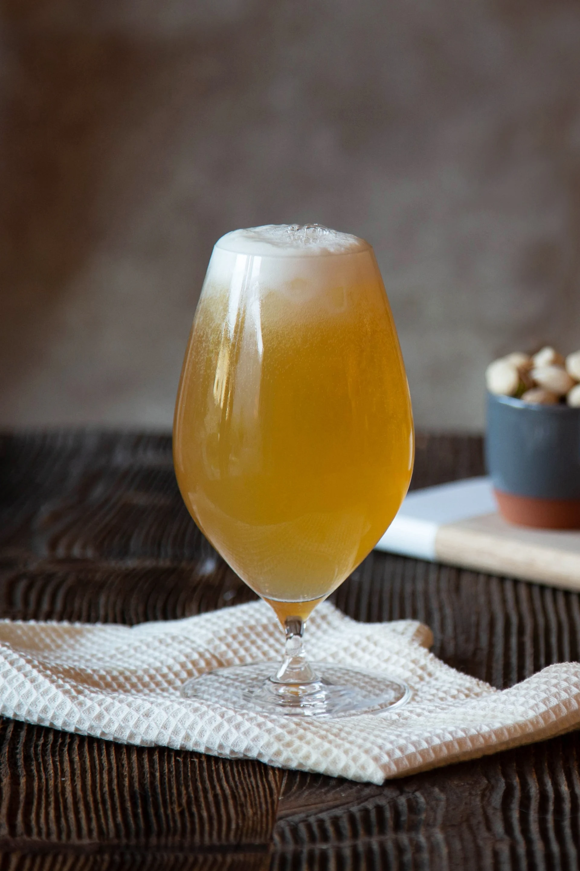 Een glas schuimig amberkleurig bier op een wit doek, met kommen met noten en grissini op de achtergrond op een houten plank.