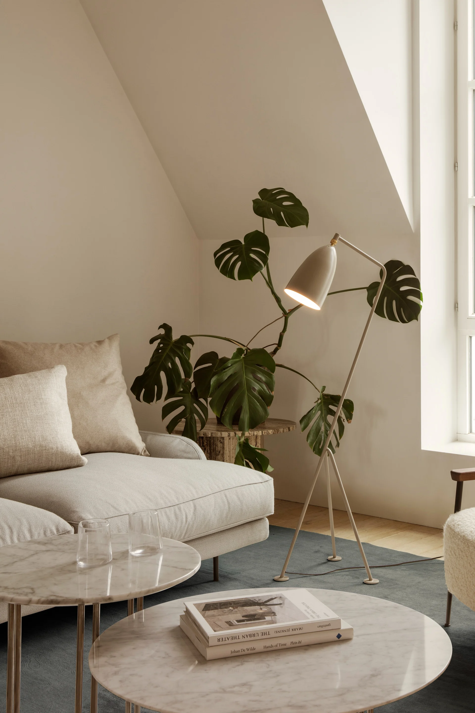 Lichte woonkamer met crèmekleurige bank, marmeren salontafels, staande lamp, plant en pluche fauteuil bij een boograam.