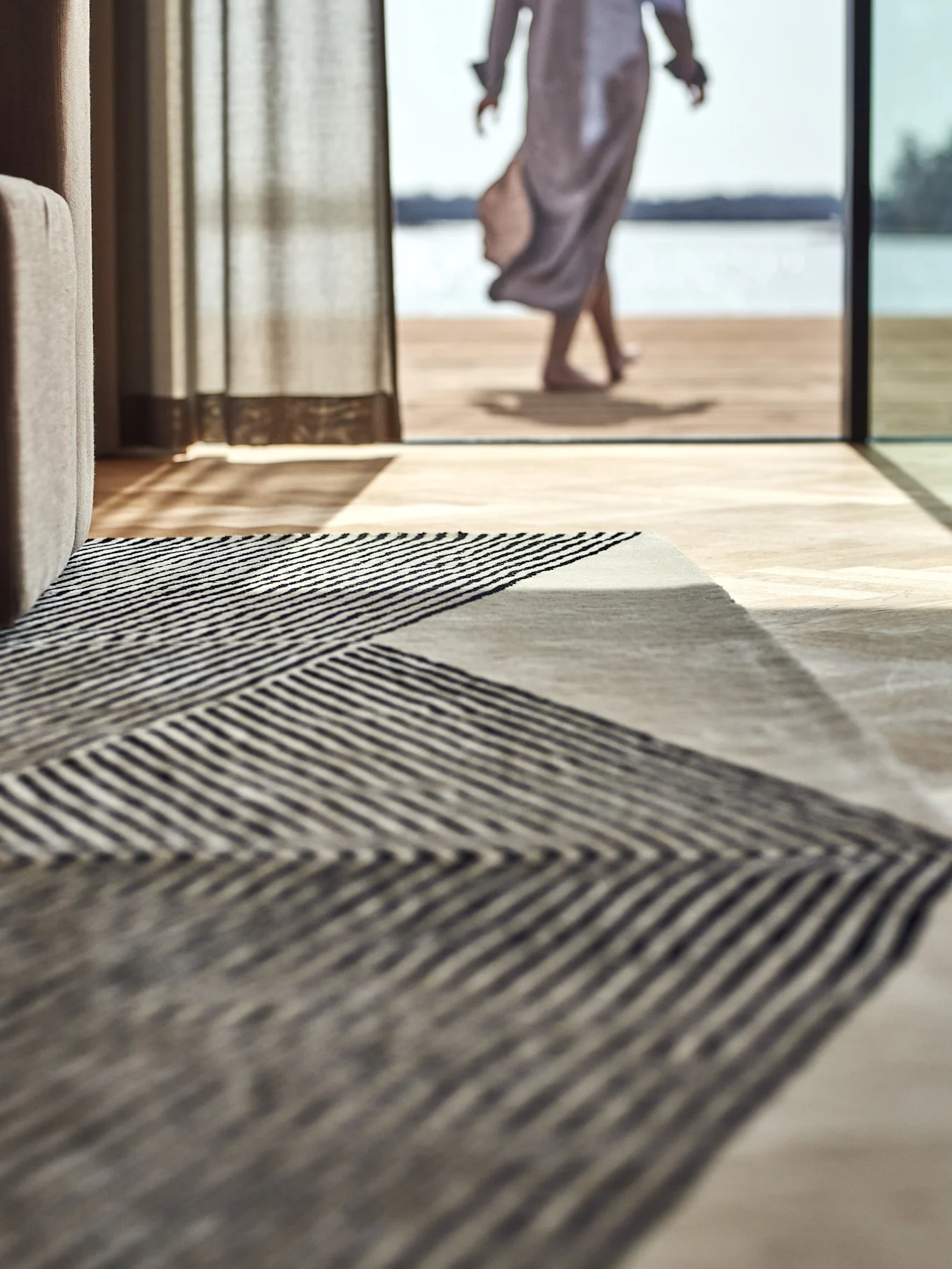 Geometrisch gestreept vloerkleed op een houten vloer, met een persoon die naar een meer buiten loopt.