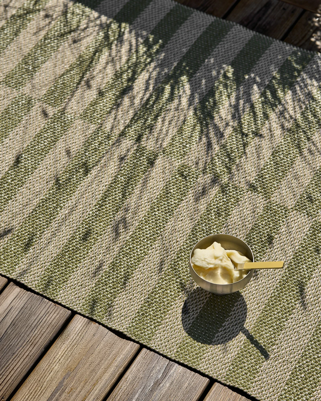 Groen en beige gestreept geweven tapijt met een kom ijs en palmenschaduwen op een houten terras.