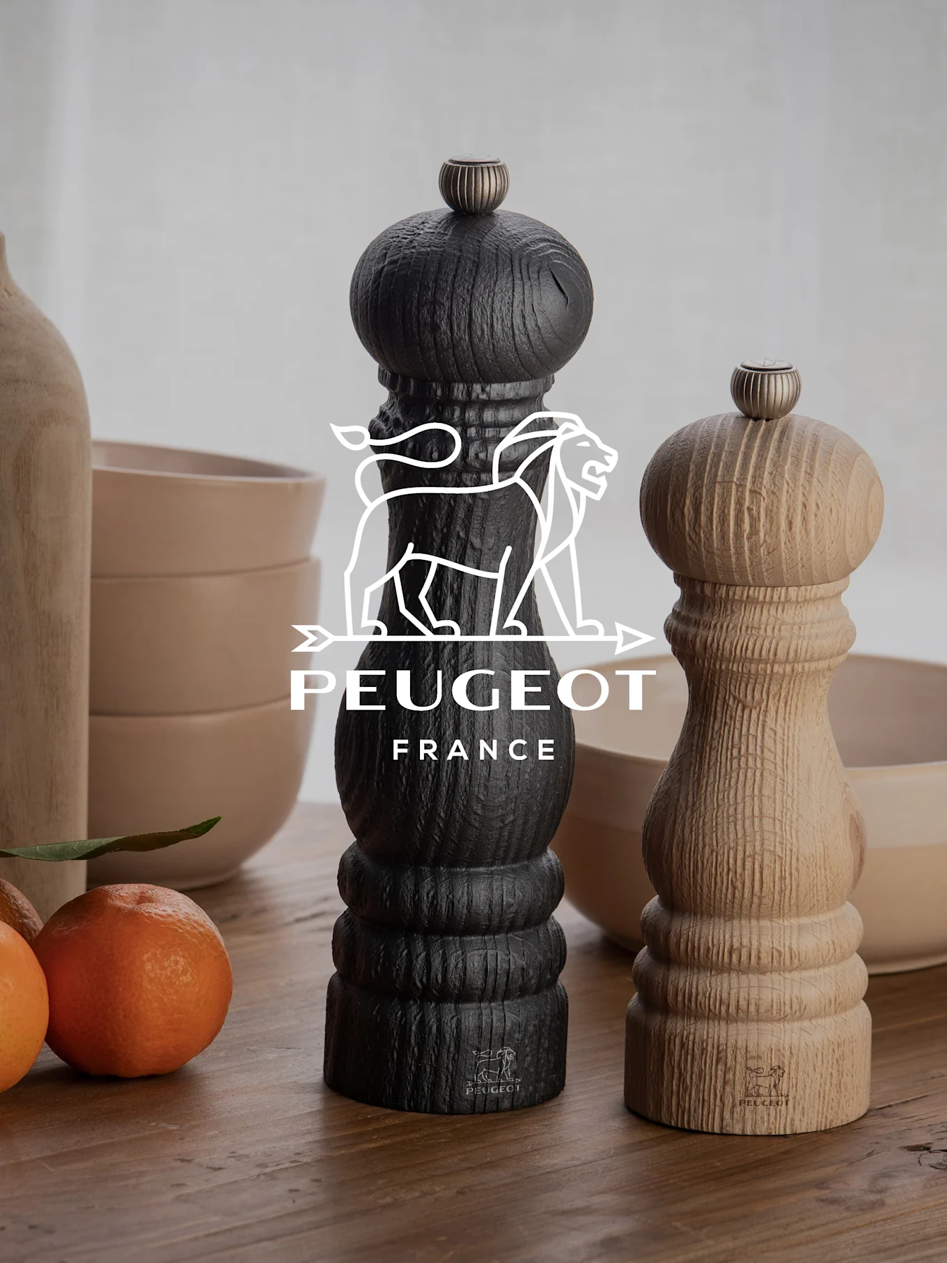 Twee Peugeot pepermolens, een zwarte en een van natuurlijk hout, op een houten tafel met mandarijnen en kommen.