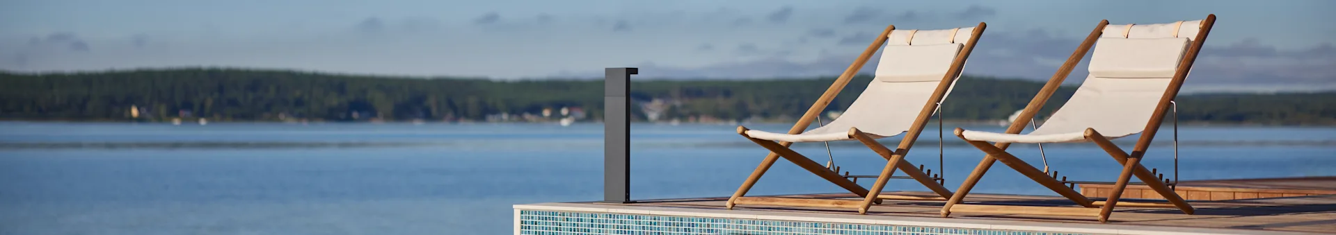 Twee witte stoffen en houten ligstoelen op een terras met uitzicht op een meer en een bos in de verte.