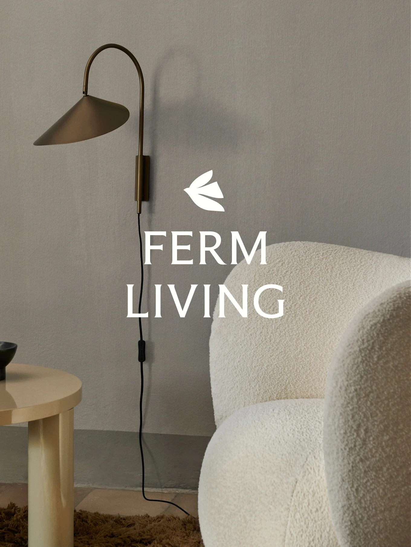 Ferm Living-logo boven een messing wandlamp, een crèmekleurige bouclé fauteuil en een lichte houten bijzettafel in een minimalistische setting.