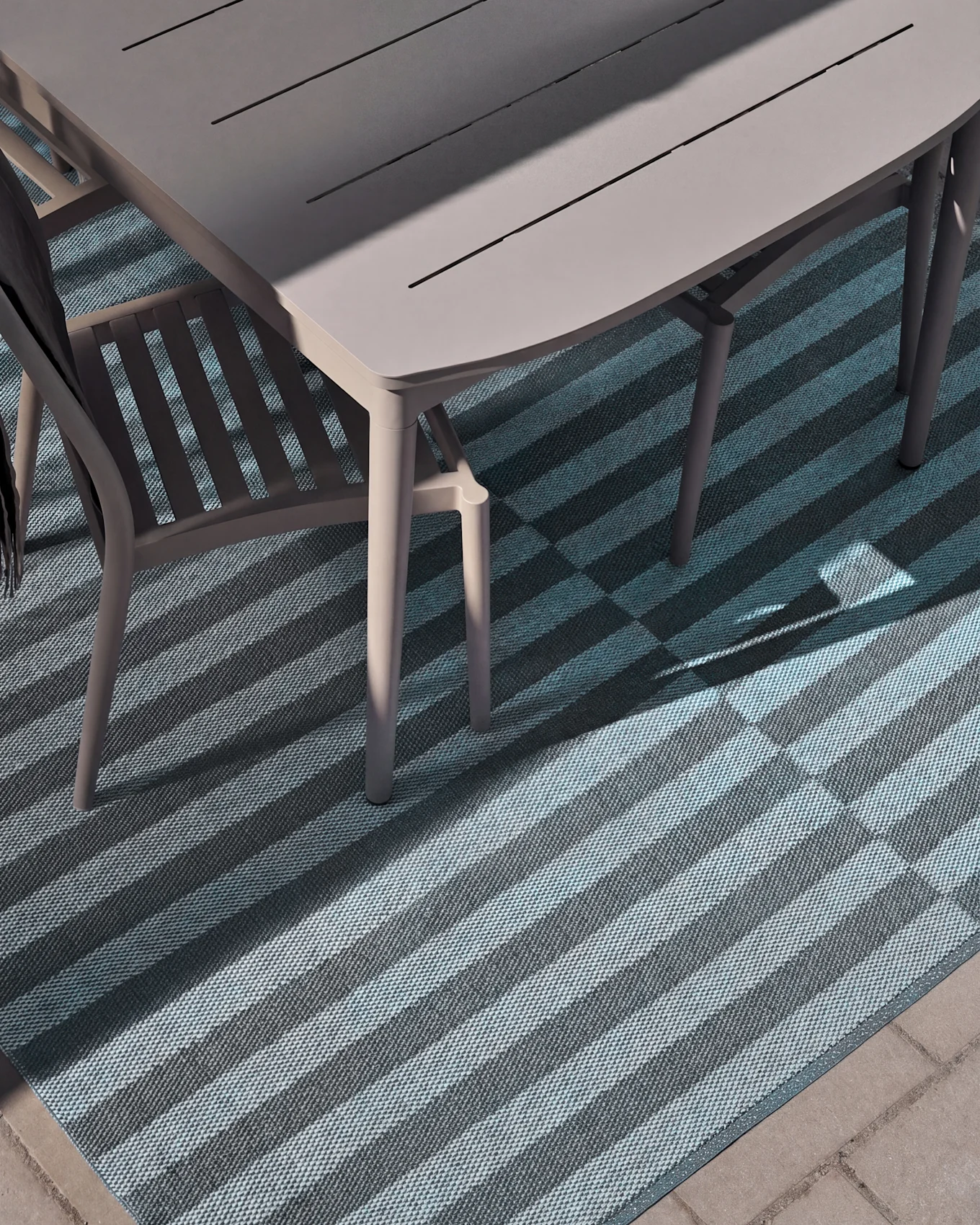 Bovenaanzicht van een buitentafel en stoel op een gestreepte mat met licht- en donkerblauwe strepen, gedeeltelijk verlicht door zonlicht.