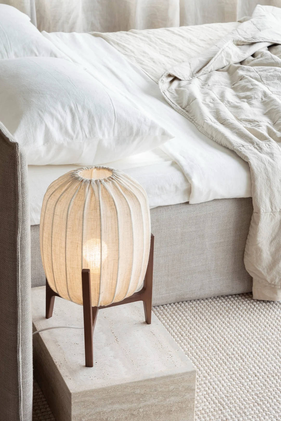 Een nachtkastje met een gloeiende geplooide stoffen lamp, naast een bed met witte lakens en een beige dekbed.