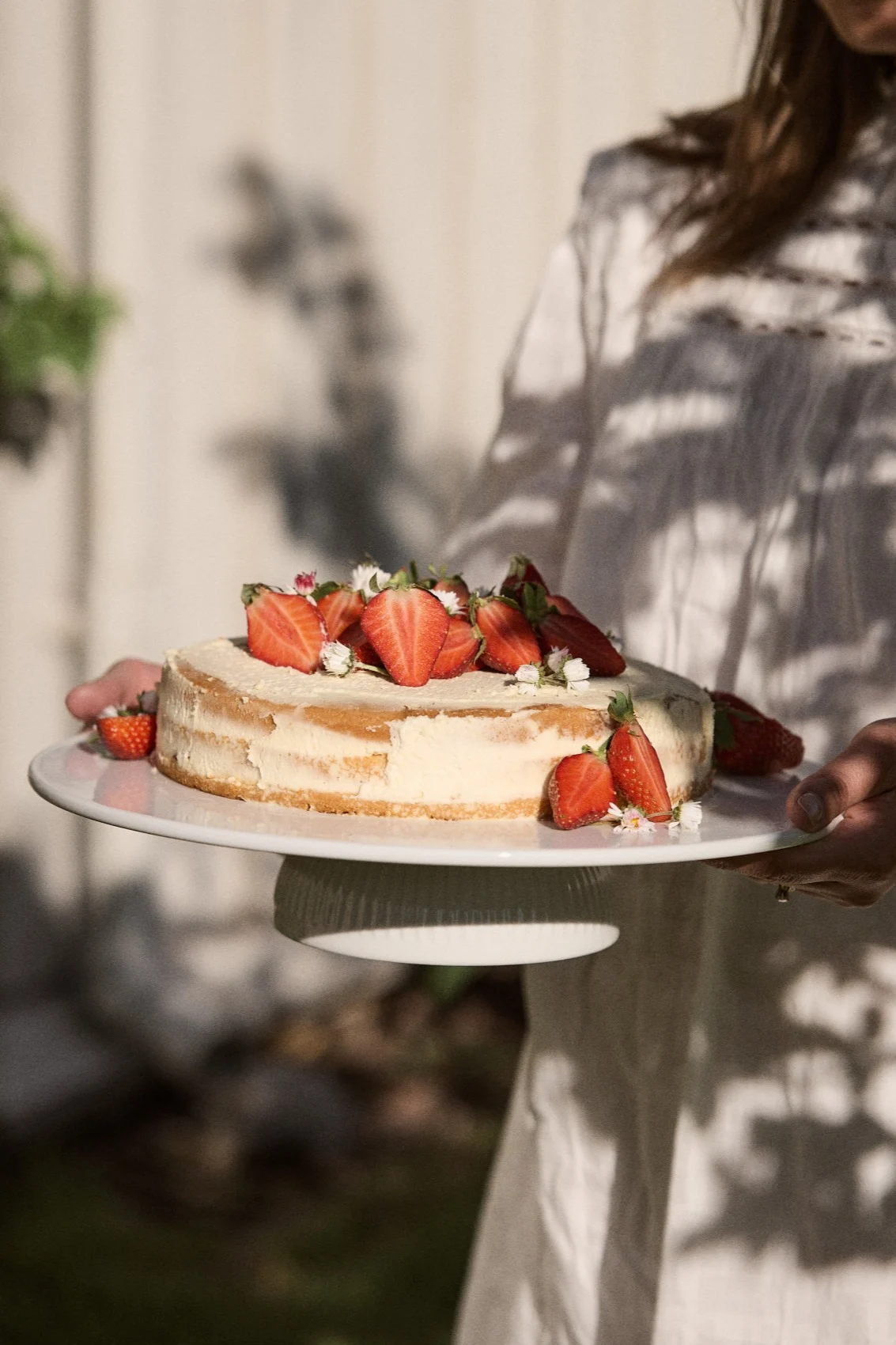 Persoon houdt een ronde taart vast, gegarneerd met verse gehalveerde aardbeien en kleine witte bloemen, op een witte cakestandaard buiten.