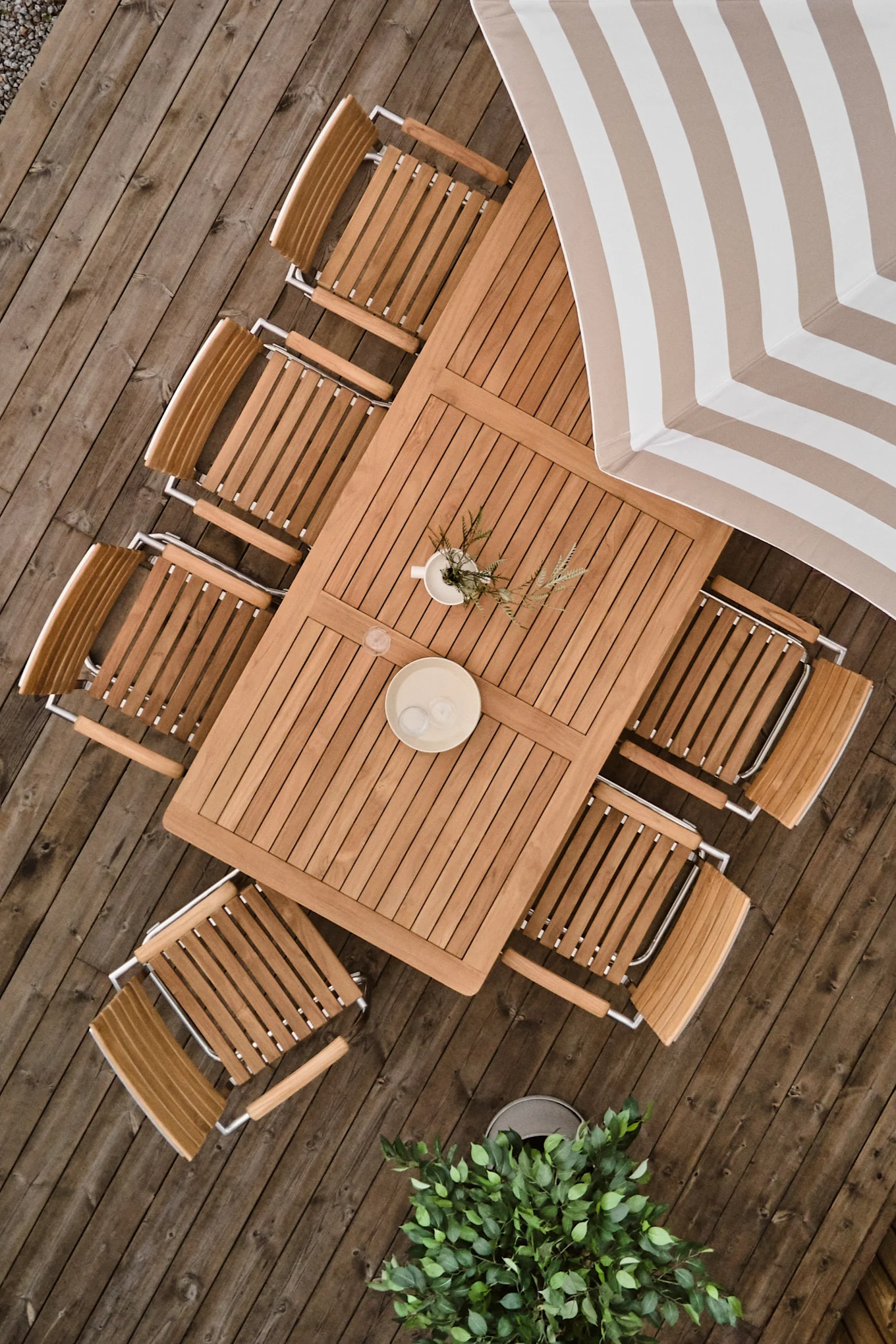 Bovenaanzicht van een rechthoekige houten eettafel met acht stoelen op een houten vlonder, een gestreepte parasol en potplanten.